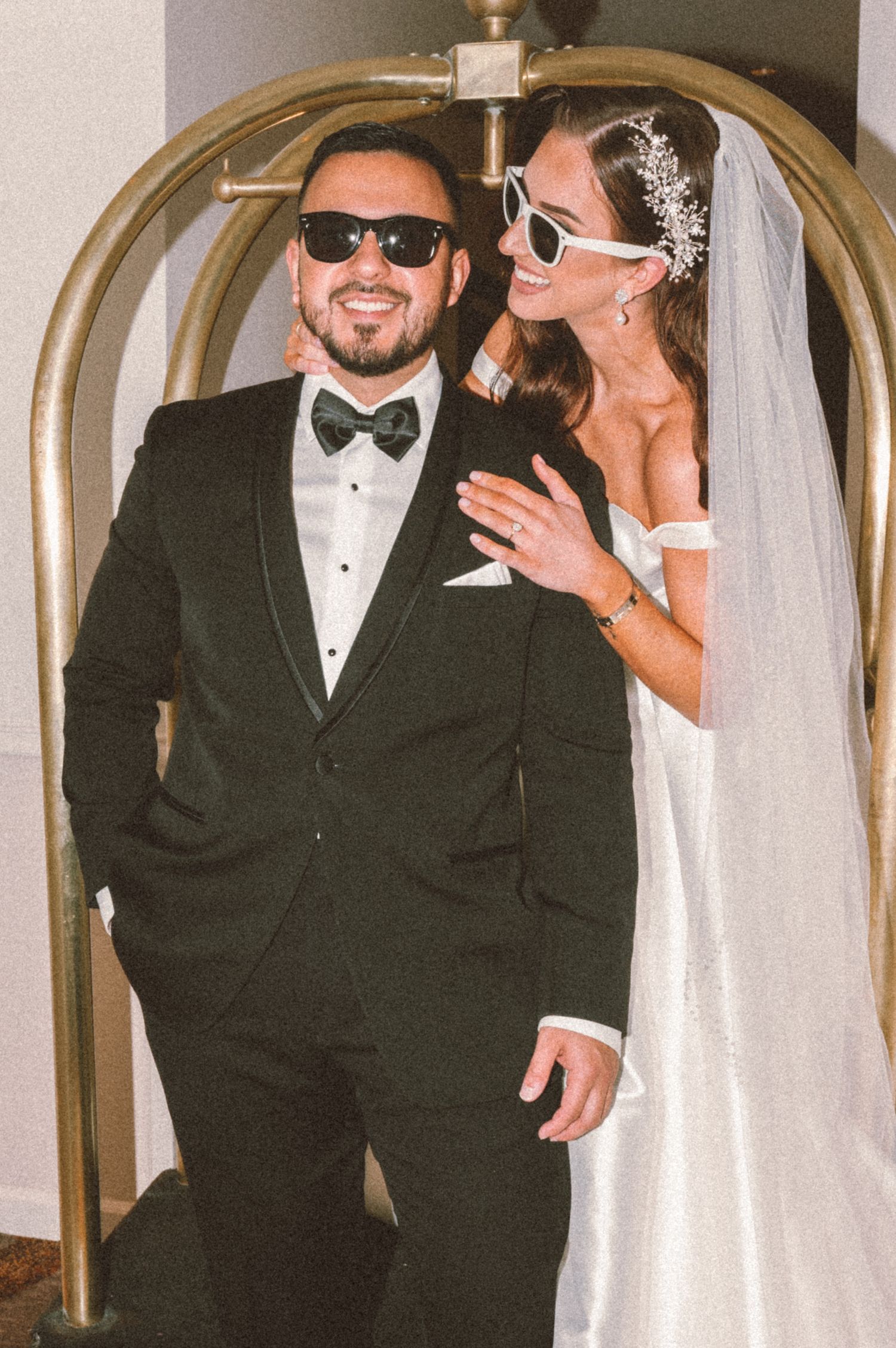 Stylish wedding couple wearing sunglasses pose under gold archway in hallway.