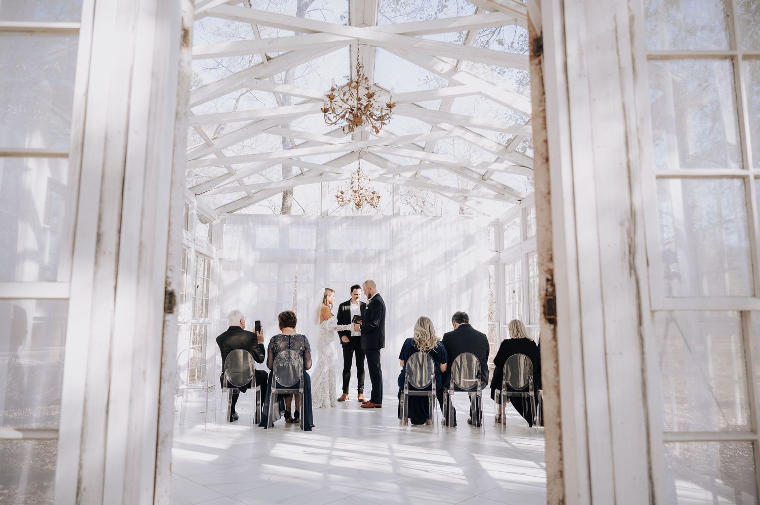 Intimate wedding ceremony in an elegant white glass greenhouse with crystal chandeliers and seated guests.