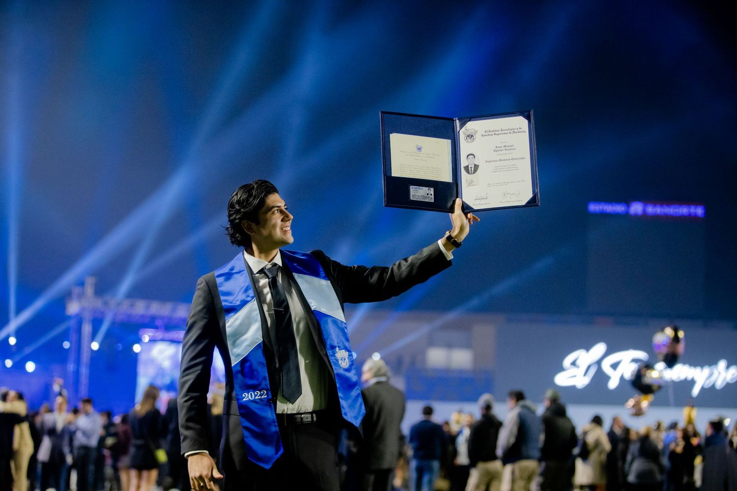 Graduate in blue sash raising diploma at a dramatic blue-lit graduation ceremony.