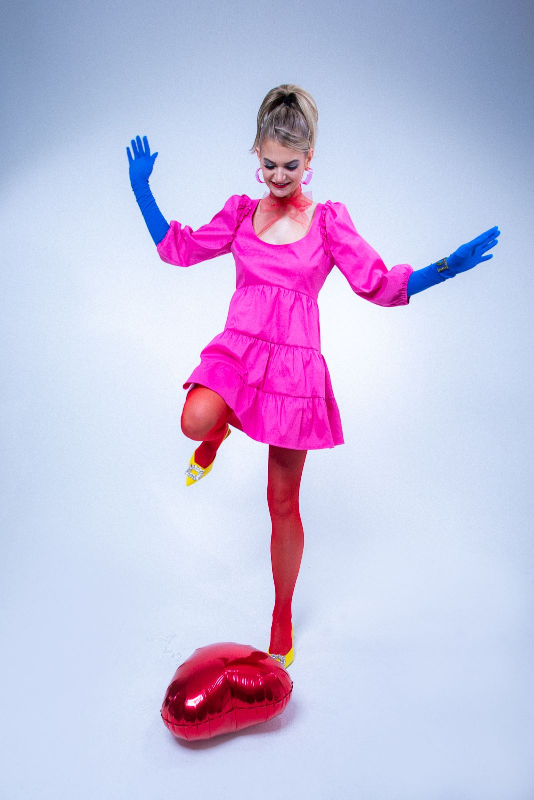 Dancer in pink dress steps on red heart balloon wearing blue gloves.