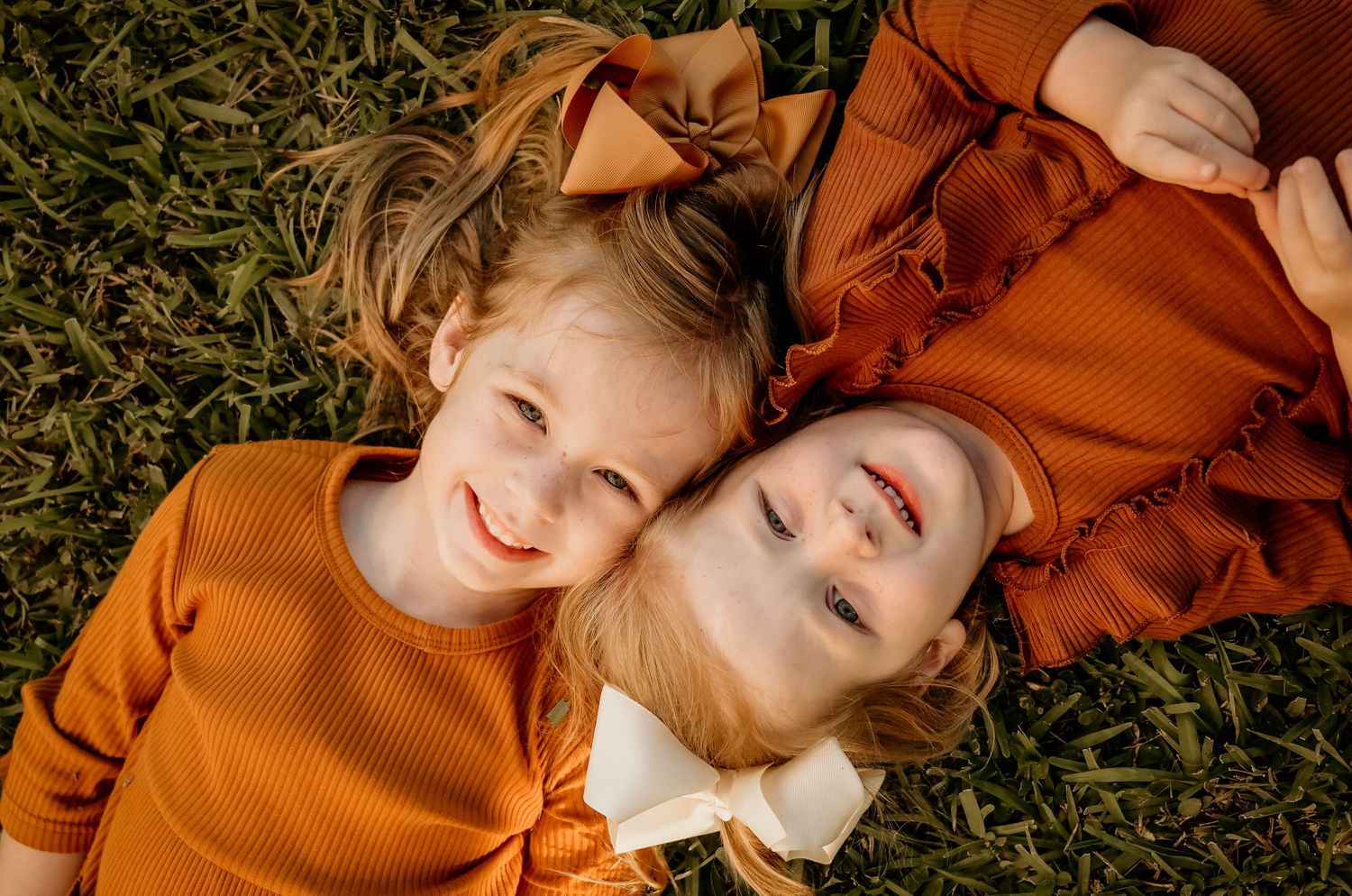 Two people wearing orange sweaters lie on grass smiling with their heads together.