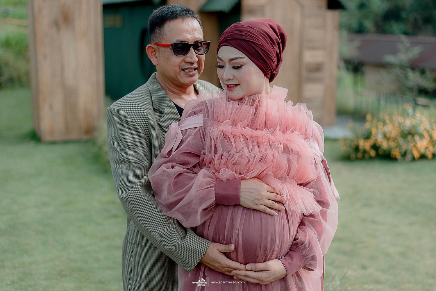 Romantic maternity photoshoot featuring a couple in pink tulle dress and grey suit in a garden setting.