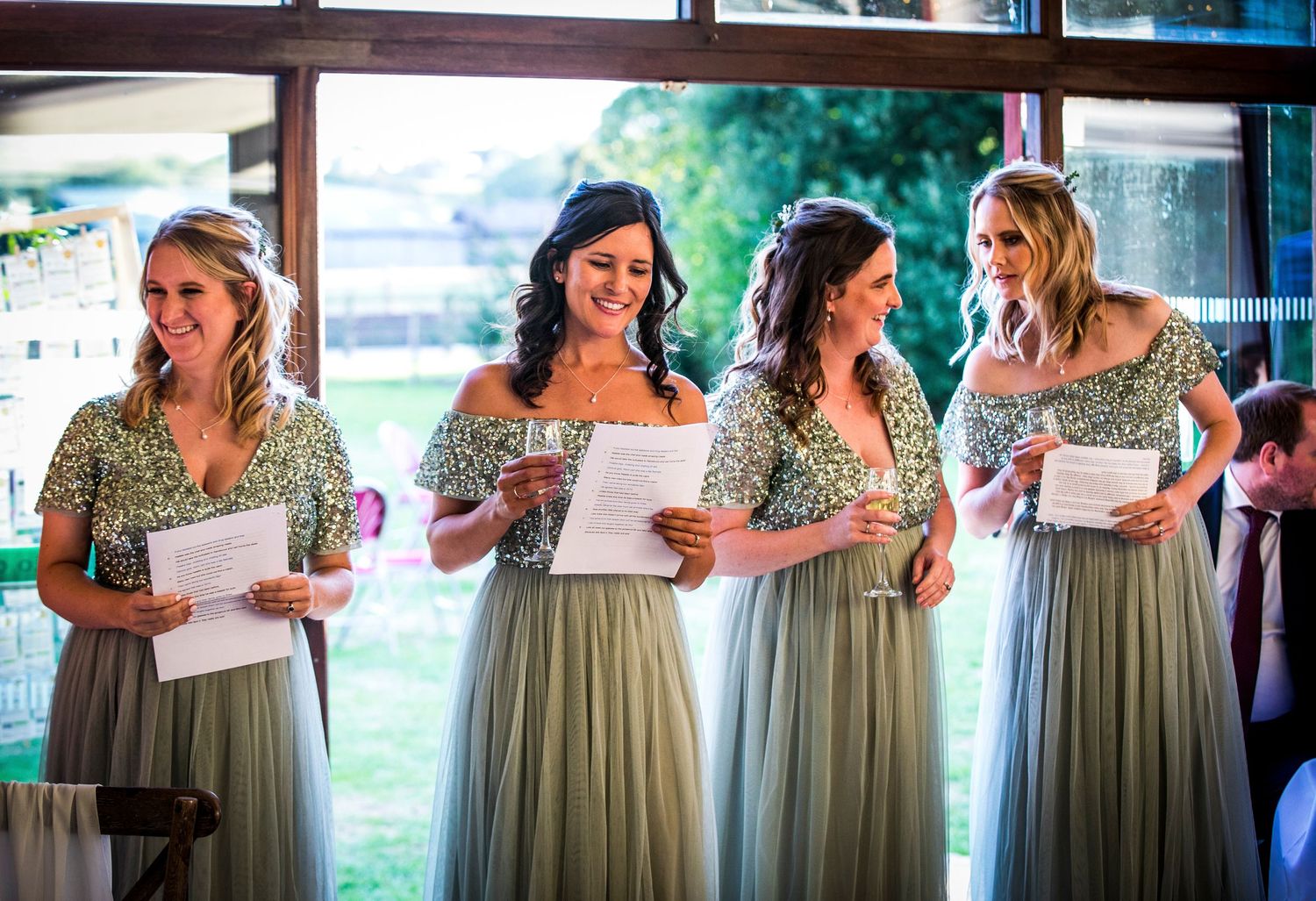 Bridesmaids in matching sage green dresses read from papers during a wedding ceremony.