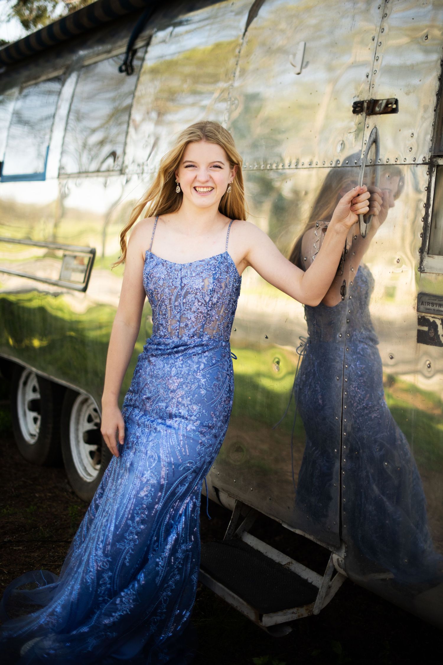 Person in sparkly blue formal dress poses against reflective metallic airstream trailer.