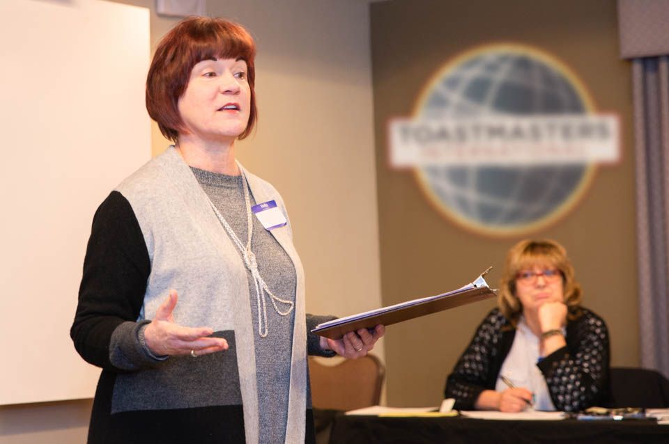 Pixs By Tina captures a woman speaking at a podium during a Toastmasters meeting with the organization's logo visible in the background.