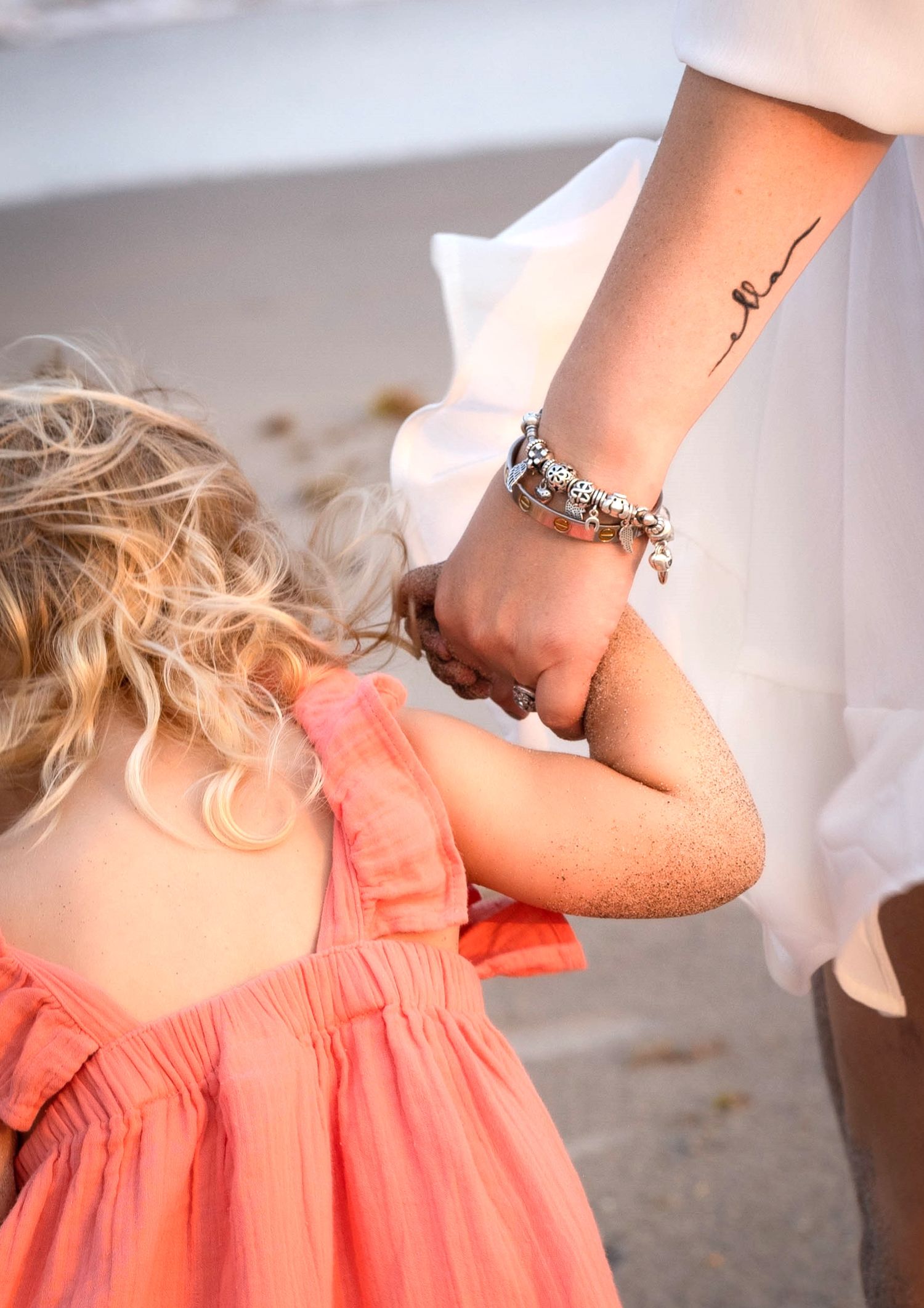 Close-up of delicate script tattoo and bracelet on wrist with coral dress in background.