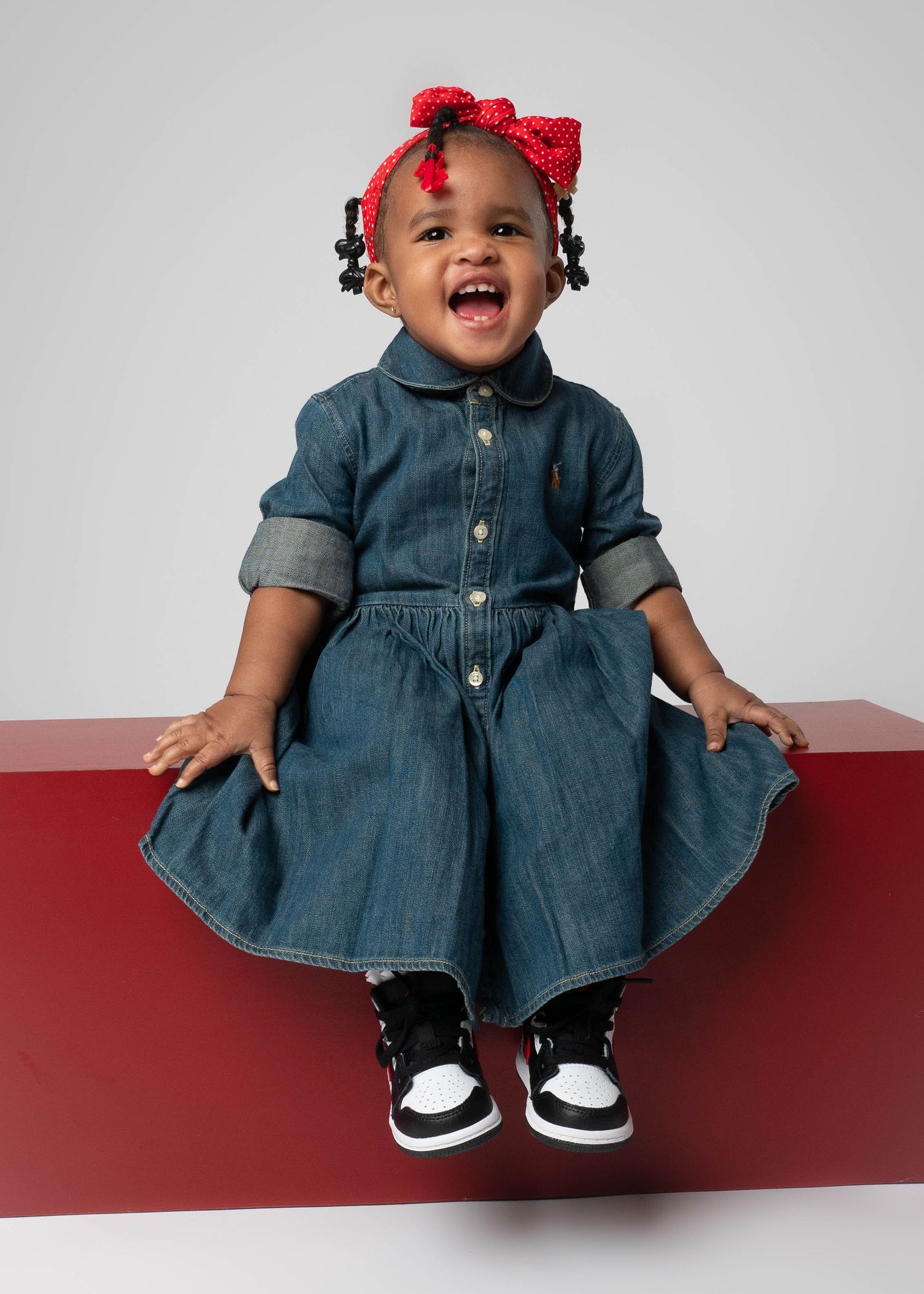 Smiling toddler in denim dress with red bow sitting on red bench.