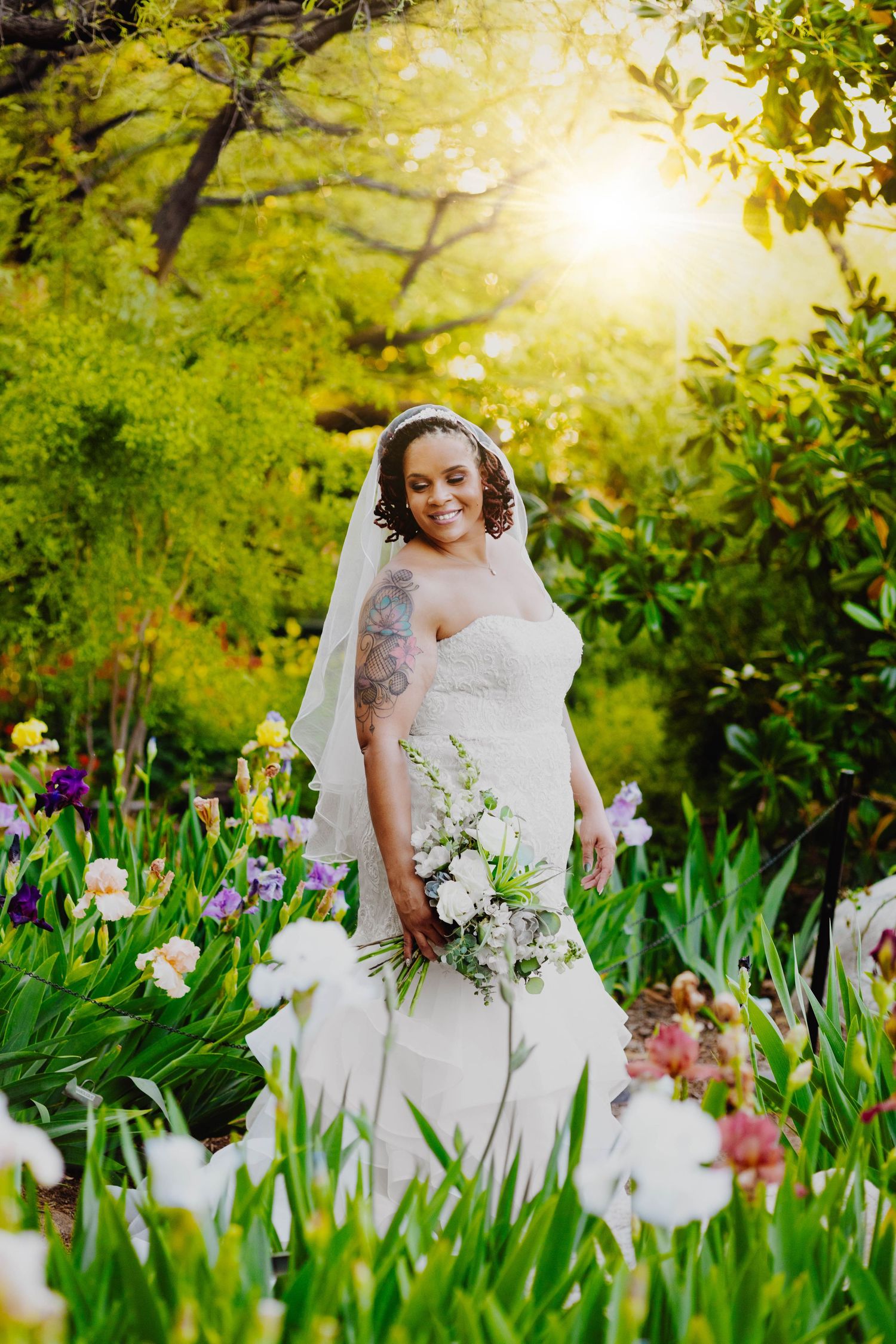 Bride in white gown stands among colorful garden flowers during golden hour.