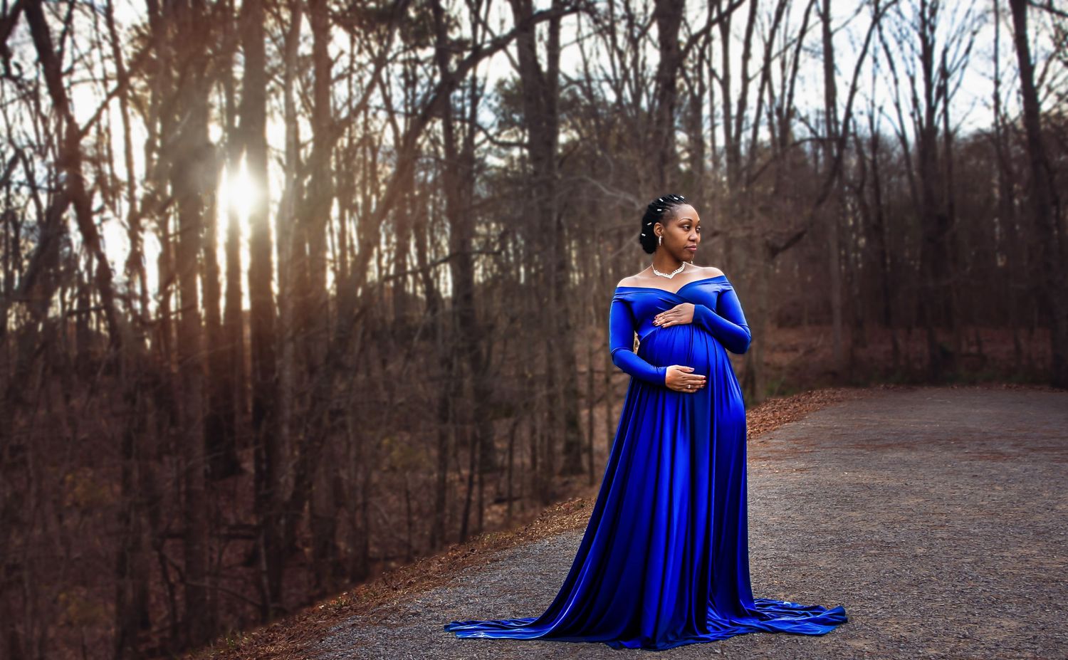 Maternity photo of woman in flowing royal blue gown against bare winter trees.