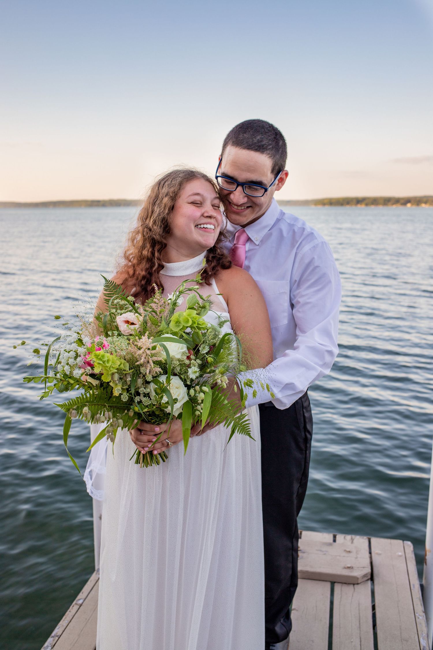 Smiling bride holding a wildflower bouquet embraced by groom on a lakeside dock at sunset.