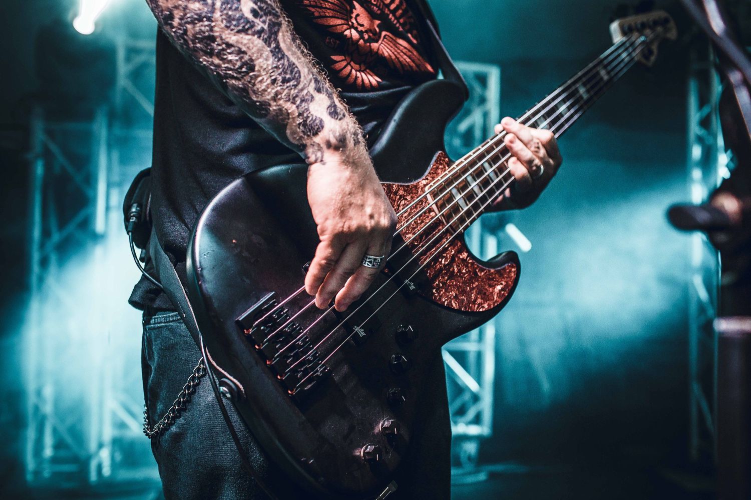 Close-up of tattooed hands playing bass guitar in blue stage lighting.