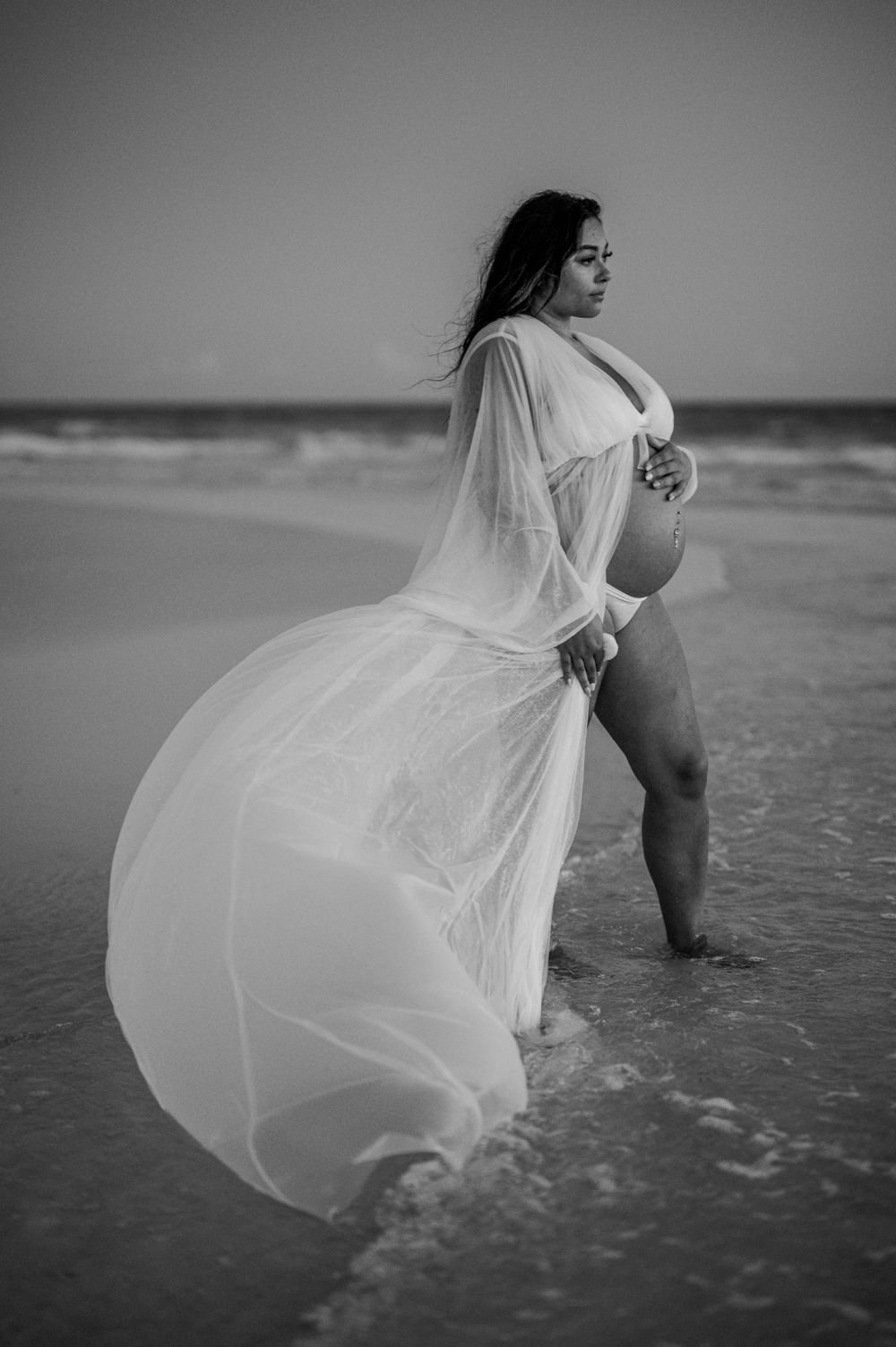Black and white beach maternity photo with billowing white dress catching ocean breeze.