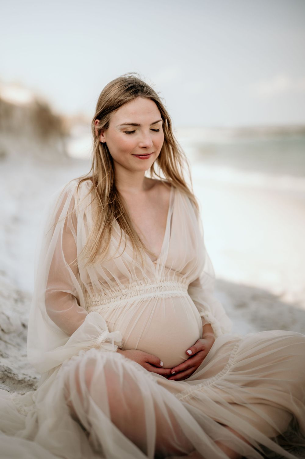 Serene maternity portrait in flowing dress against soft beach background.