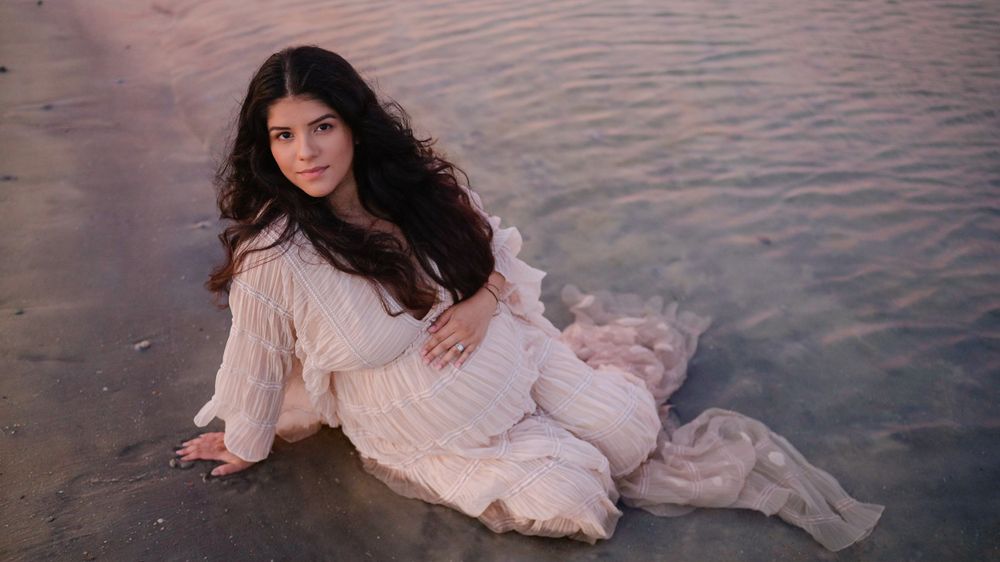 Artistic maternity portrait of woman reclining by the water in a light cream dress.