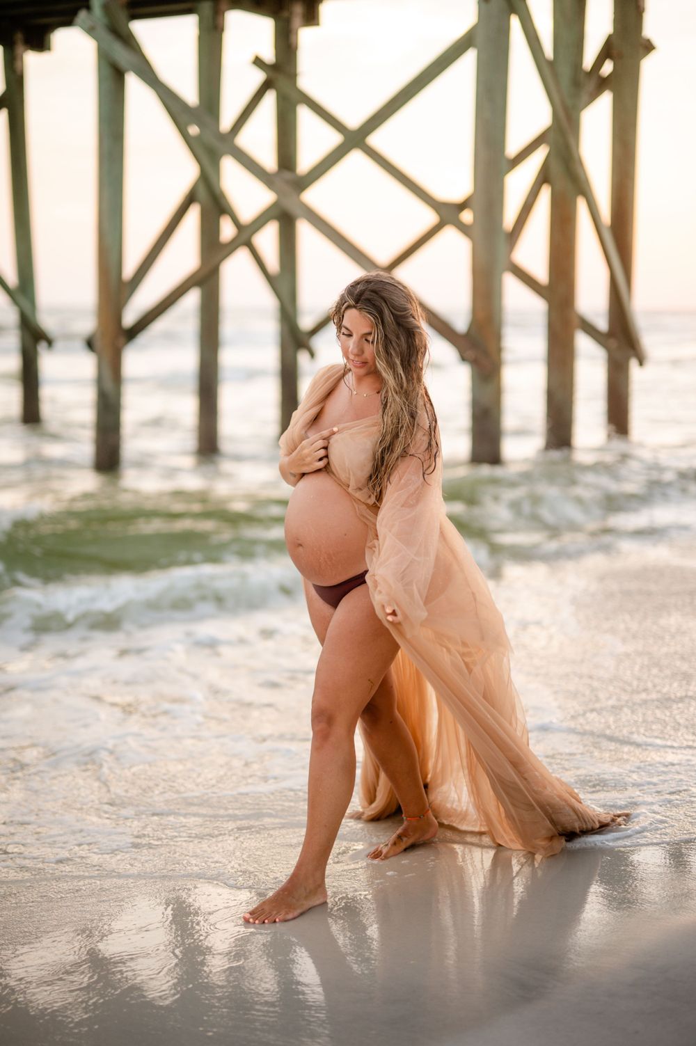 A maternity photo shoot at sunset by a wooden pier with flowing sheer peach dress and waves in the background.