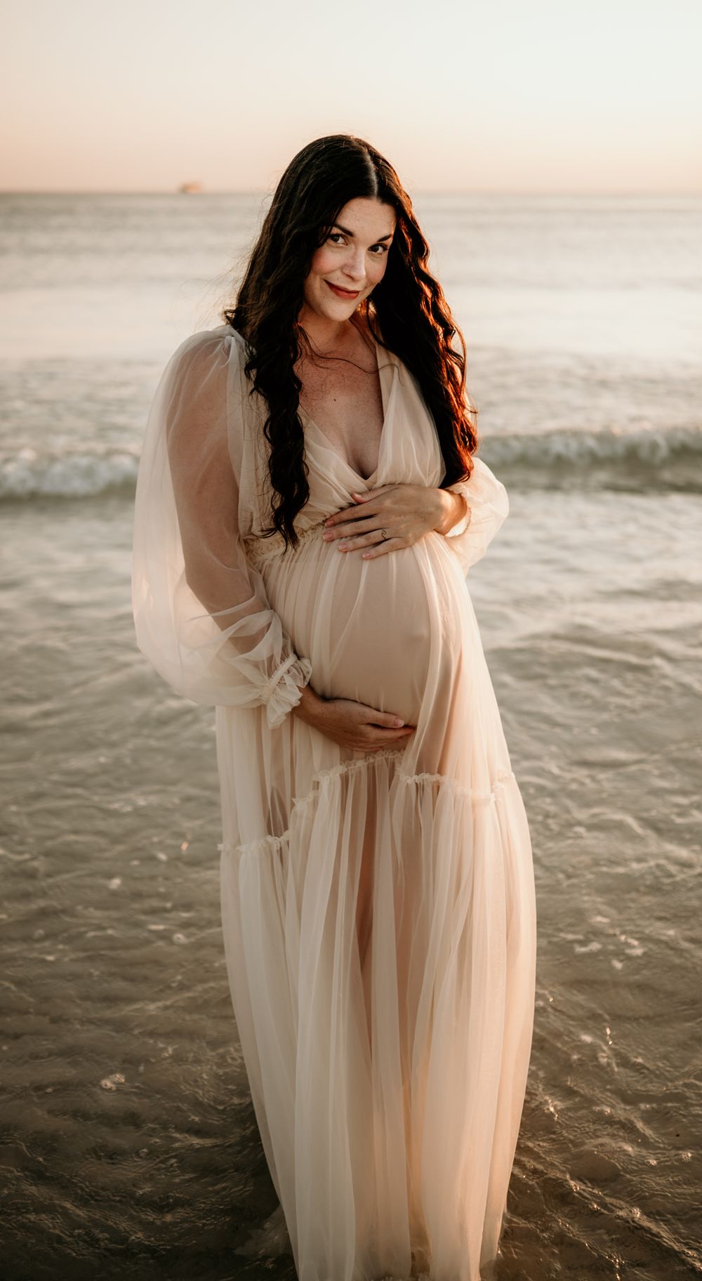 Elegant maternity portrait on beach at sunset with flowing sheer dress.