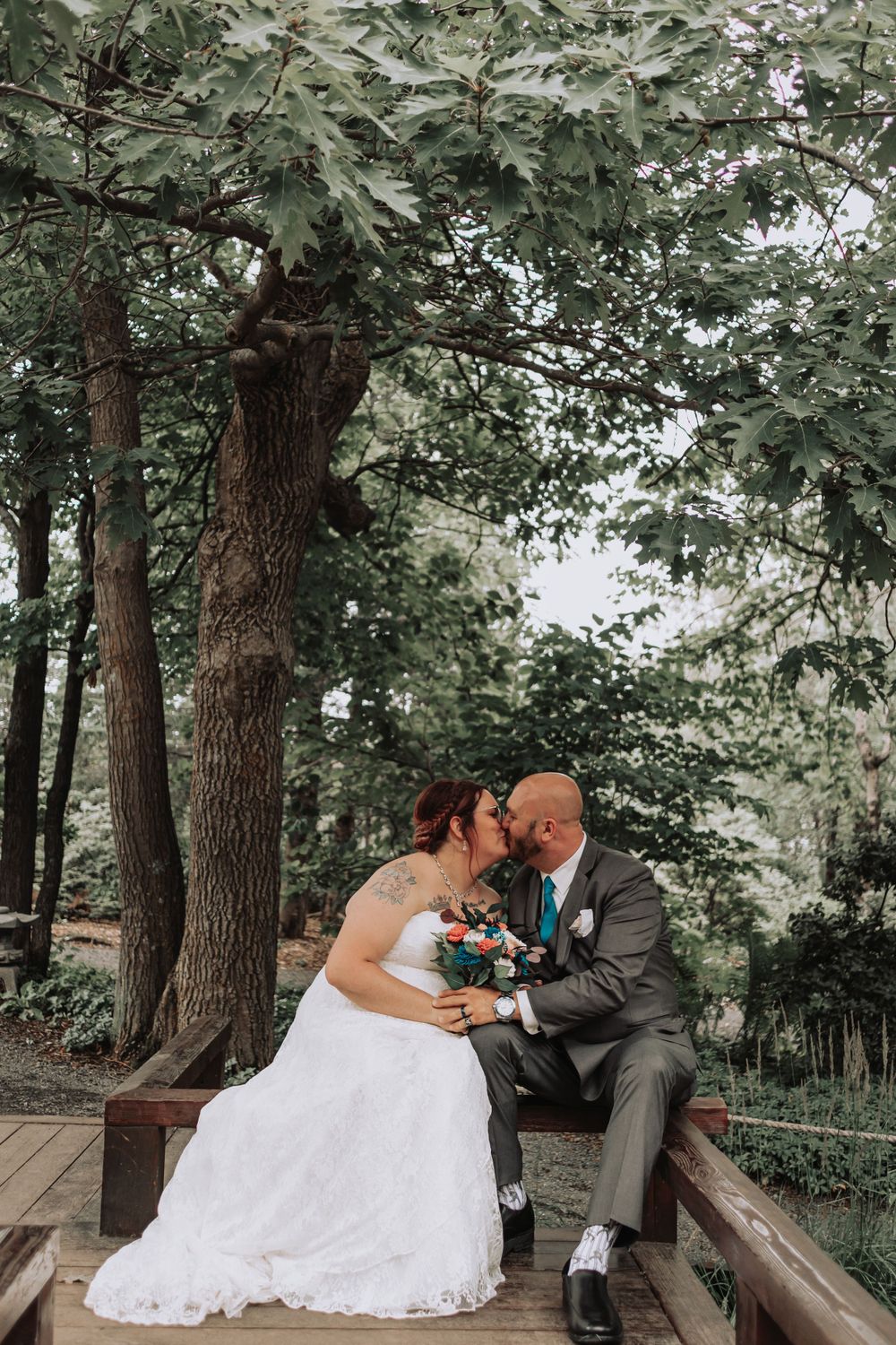A couple shares their first kiss as newlyweds on a wooden bridge surrounded by lush trees.