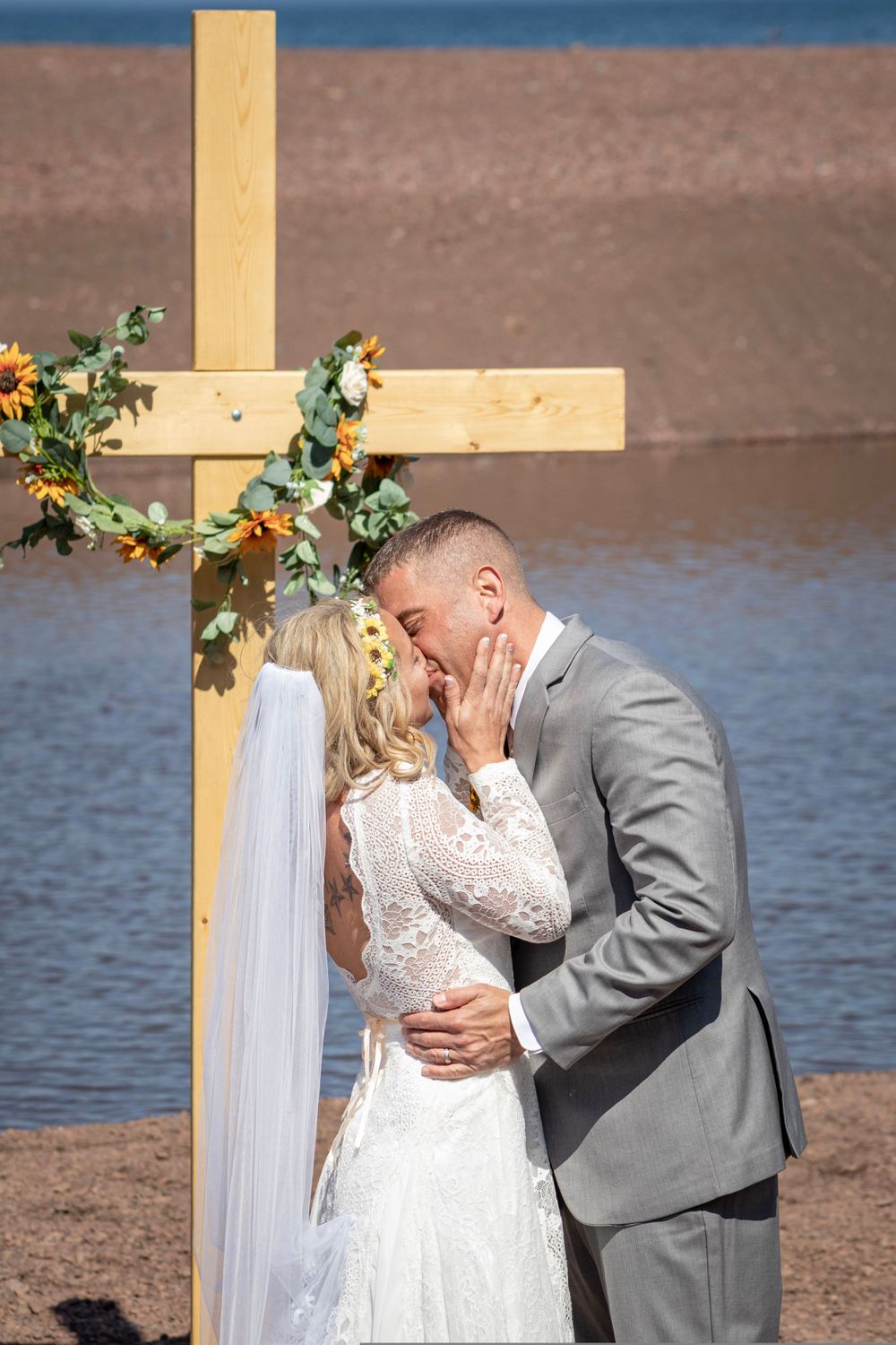 A romantic wedding ceremony moment captured by a lake with a wooden cross and floral decorations.