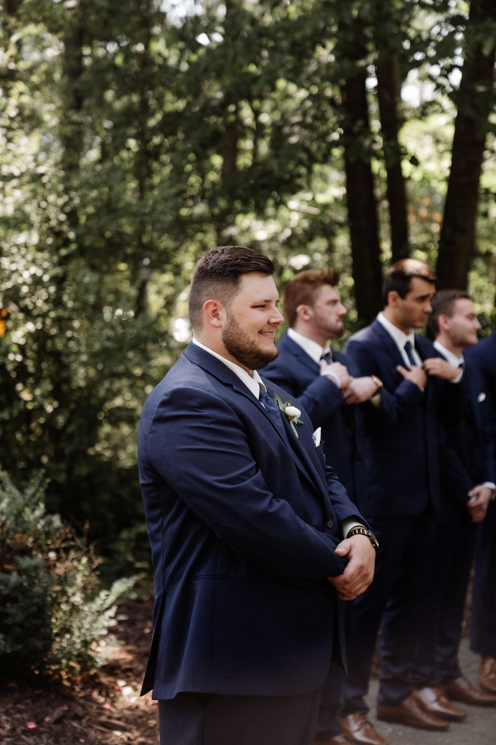 Groomsmen in navy suits showing emotional reactions during an outdoor wedding ceremony.