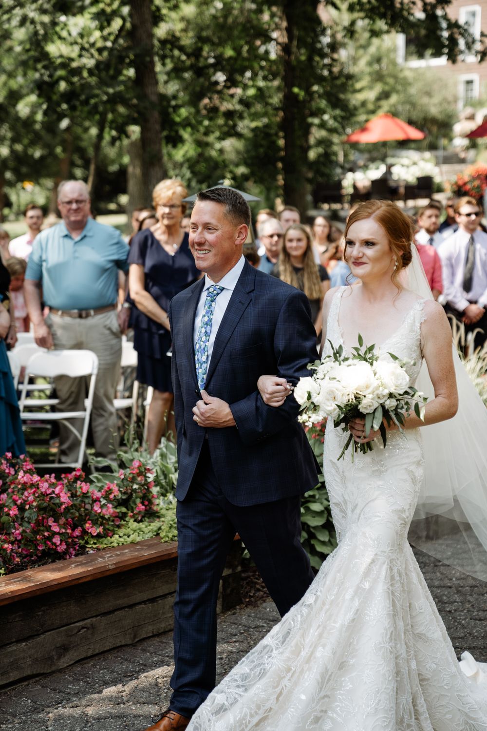 A bride in a mermaid wedding dress walks down an outdoor aisle with her escort during a garden ceremony.