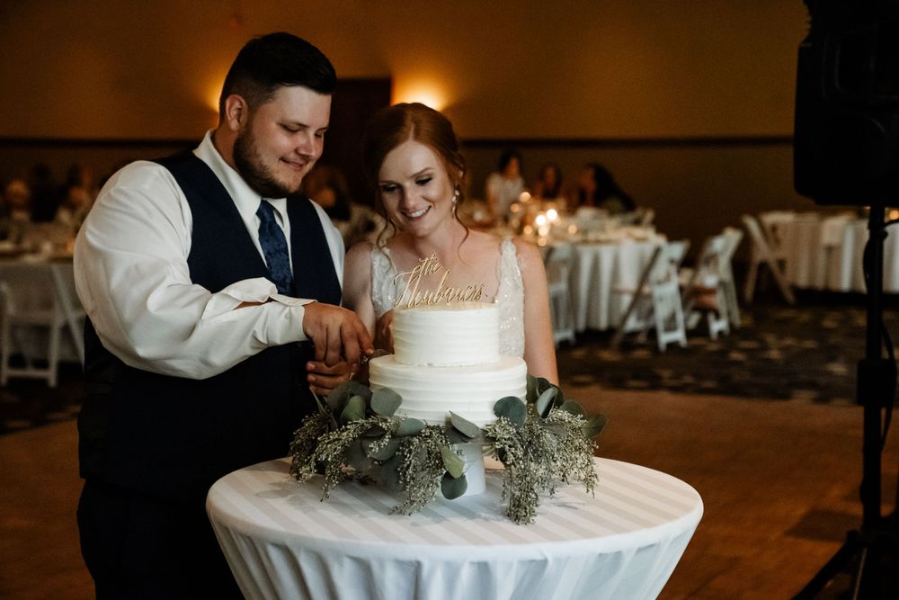 A couple cuts their elegant white wedding cake decorated with greenery at their reception.