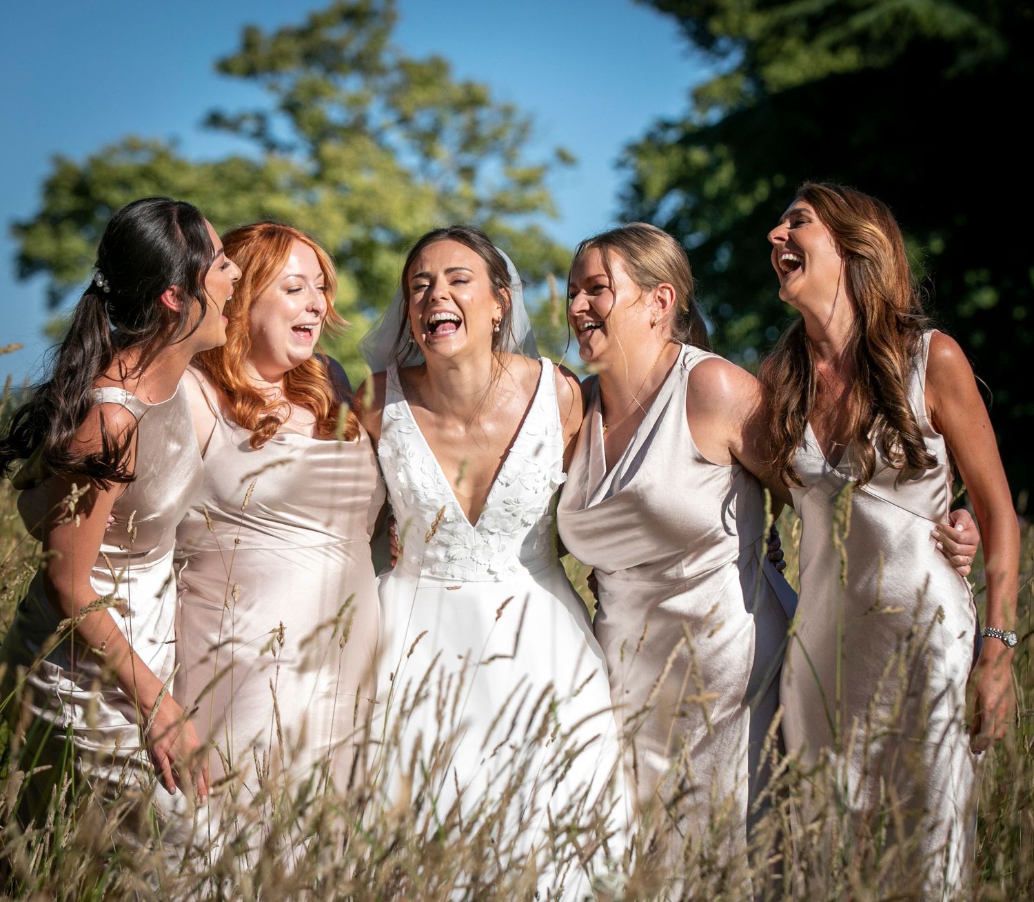 Bridesmaids in white dresses laugh together while standing in tall grass on a sunny day.