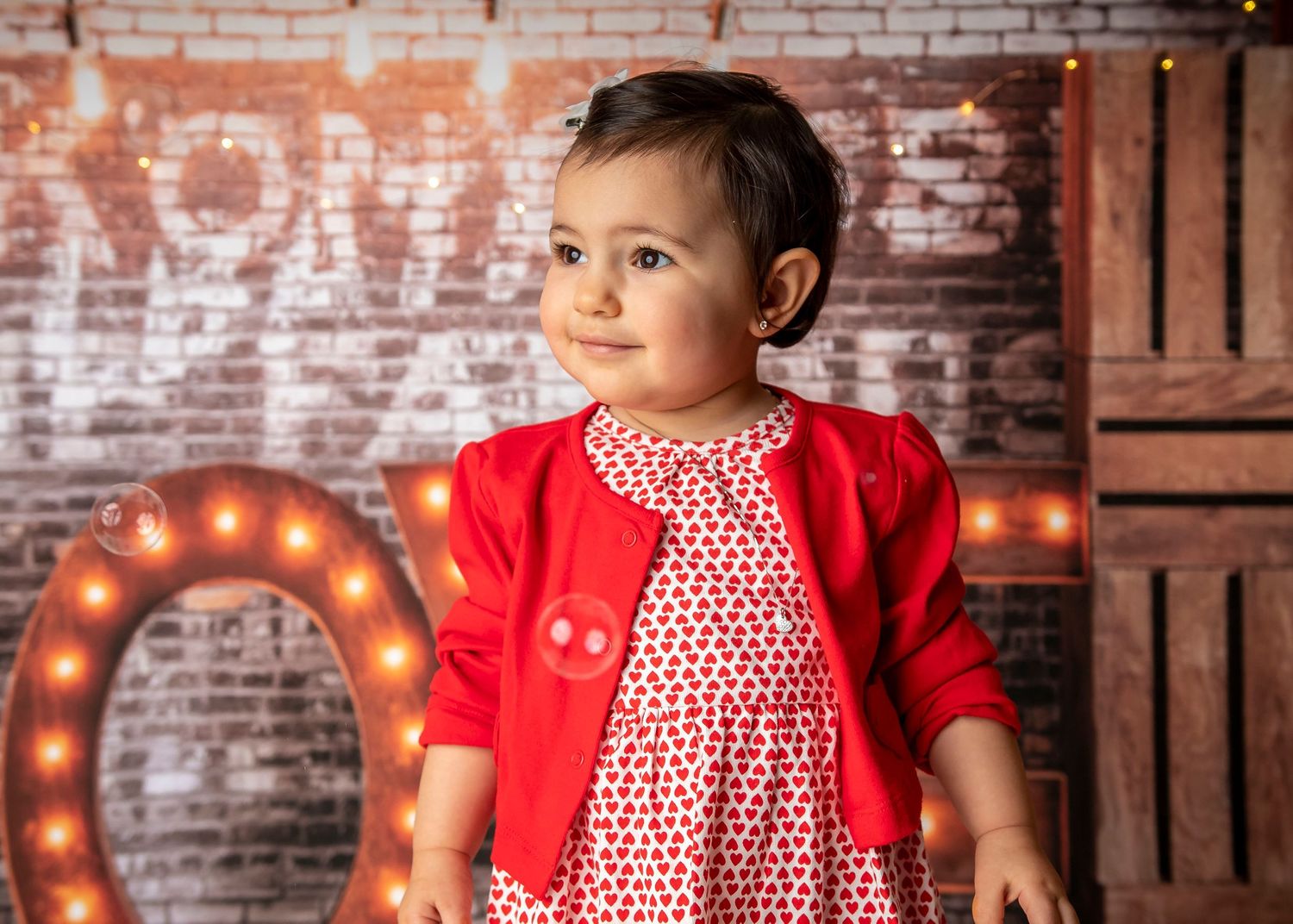Child wearing red cardigan and patterned dress poses by brick wall with vintage LOVE sign.