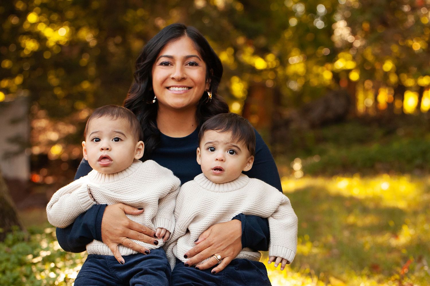 Parent holding two young babies dressed in white sweaters with fall colors in background.