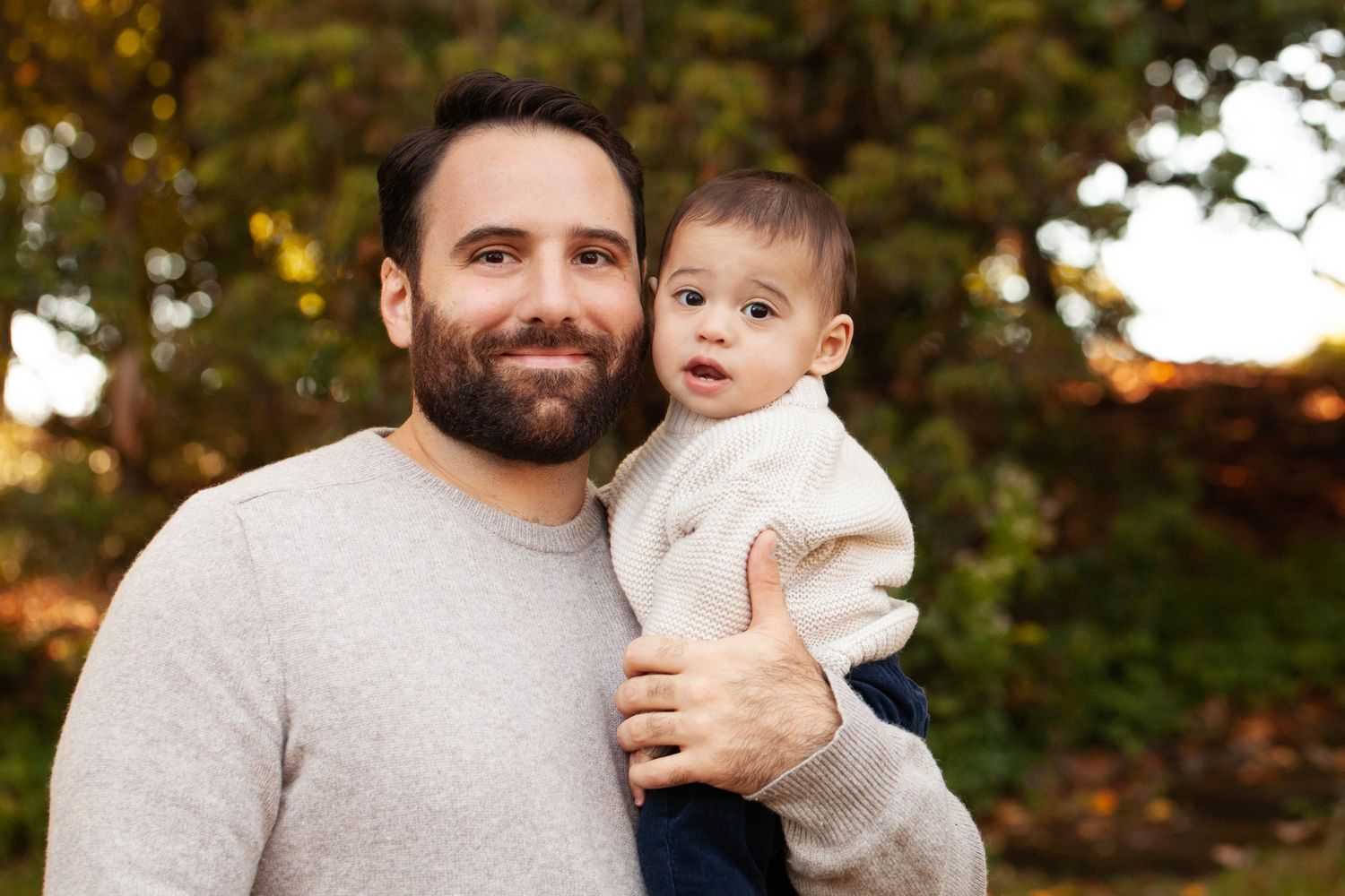 Portrait of a parent and young child in matching cream sweaters against autumn foliage.