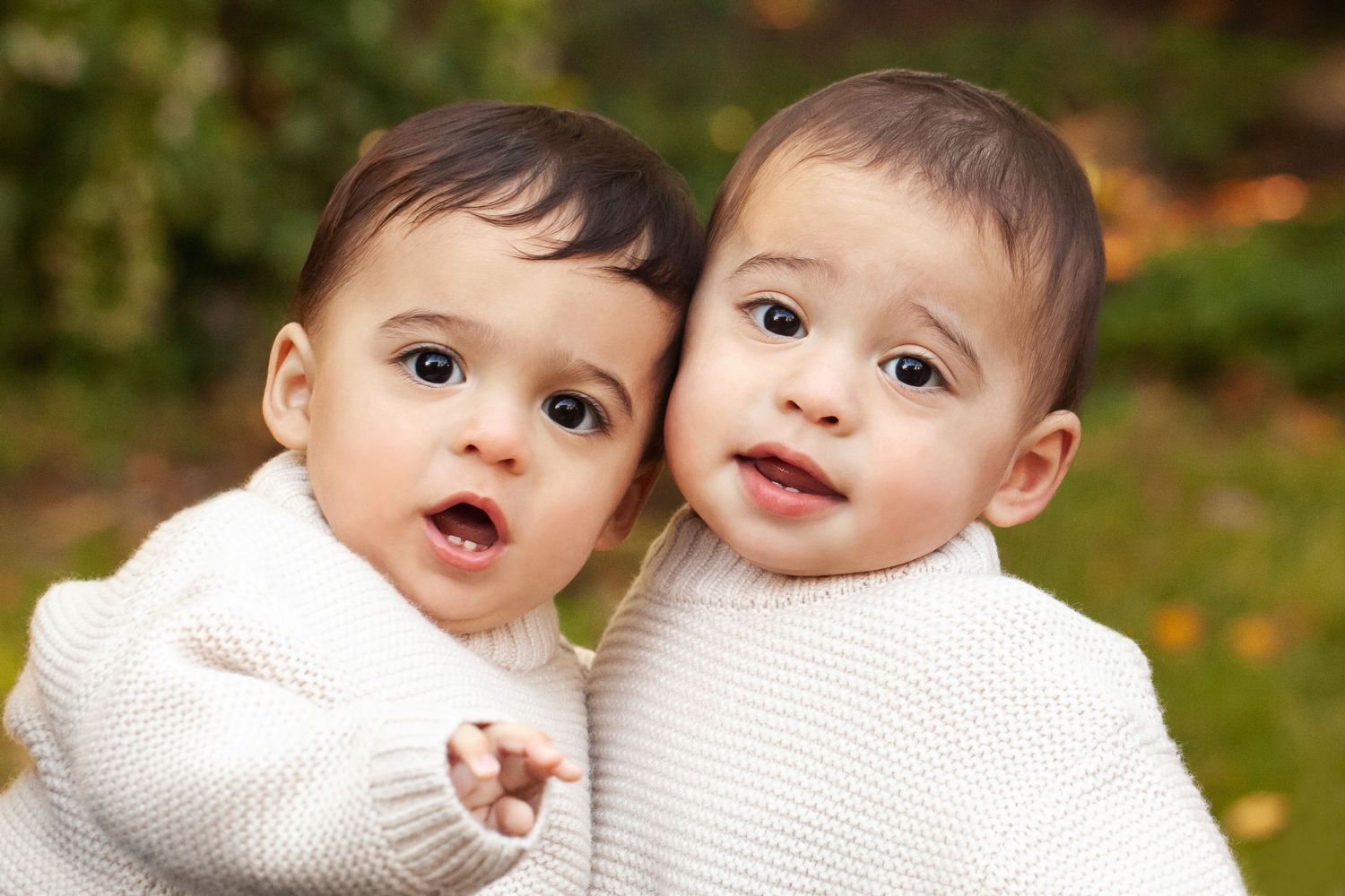 Two babies in matching white knit sweaters smiling at camera outdoors.