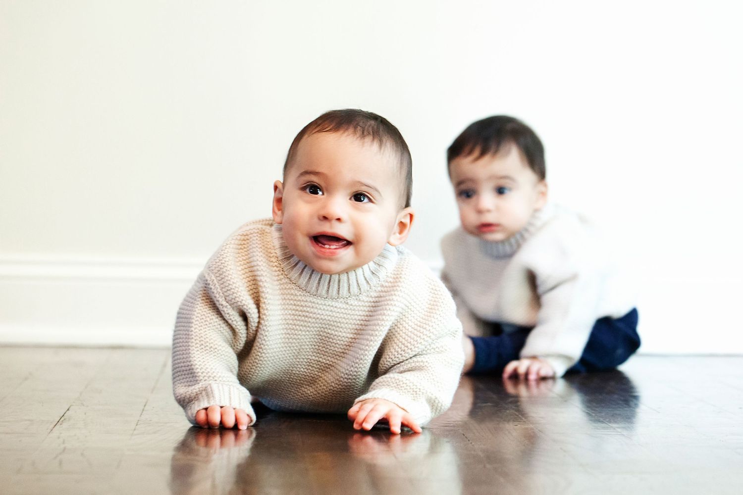 Two babies in cream sweaters crawling on hardwood floor indoors.