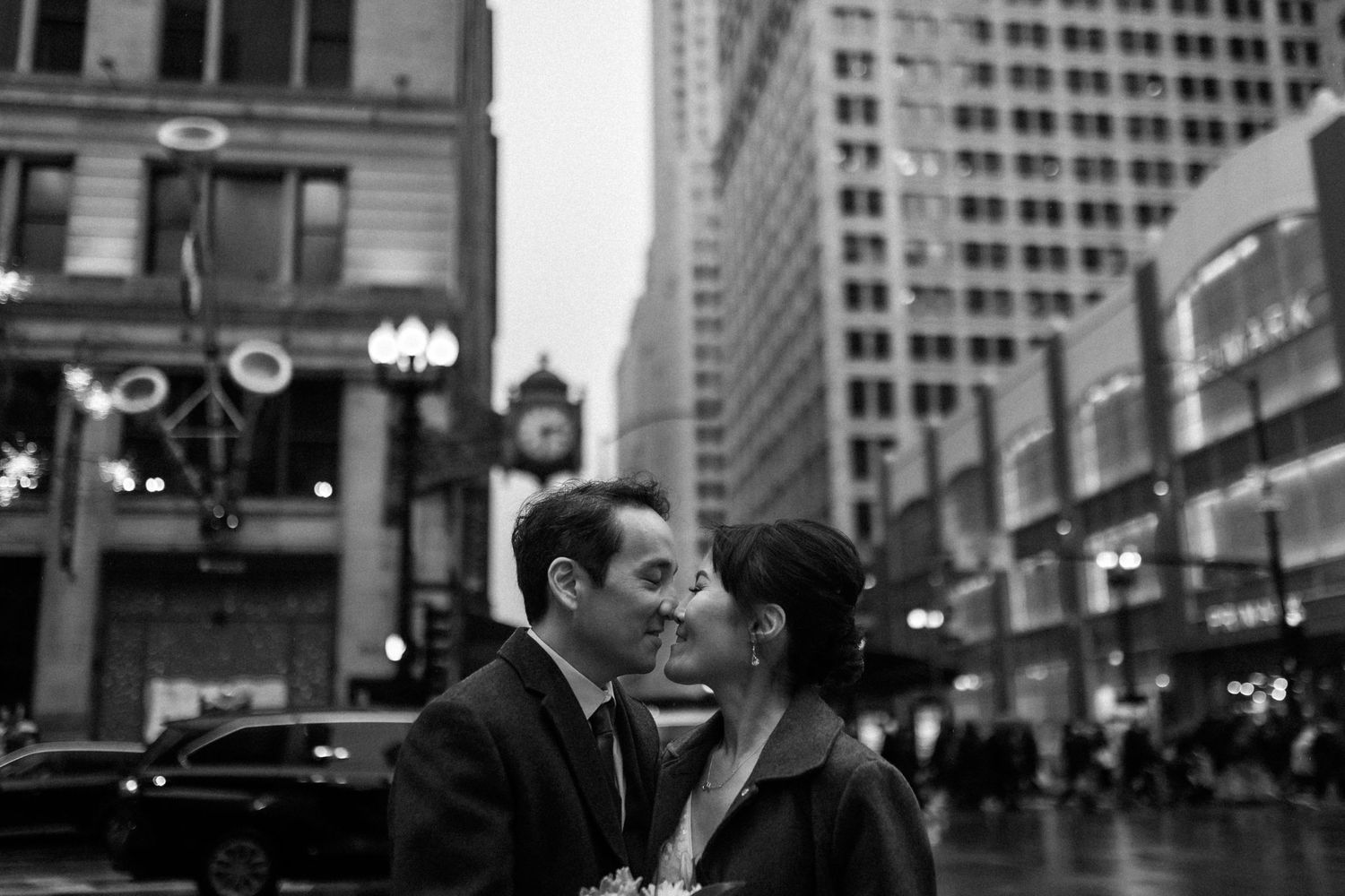 A couple shares an intimate moment on a busy city street in black and white.