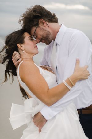 Wind-swept romantic embrace on cloudy day with couple in formal white attire in dramatic and cinematic photography taken in Hilton Head.