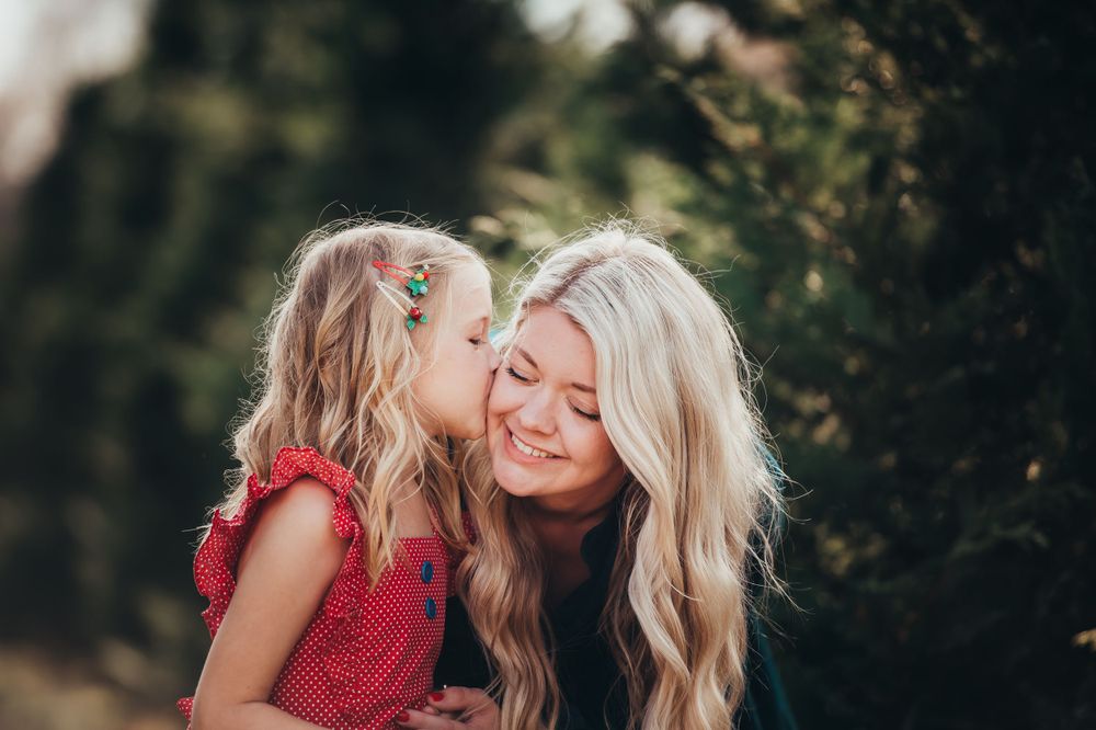 little blonde girl kisses her blonde mom on the cheek at Christmas tree farm for mini session