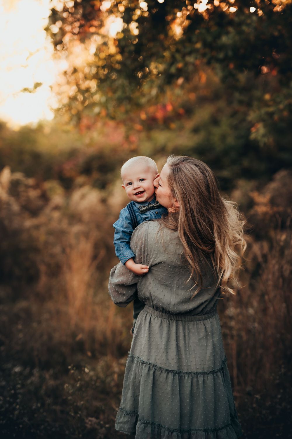 Mother's back is turned with smiling baby boy held up over her shoulder as she turns to kiss his cheek in fall