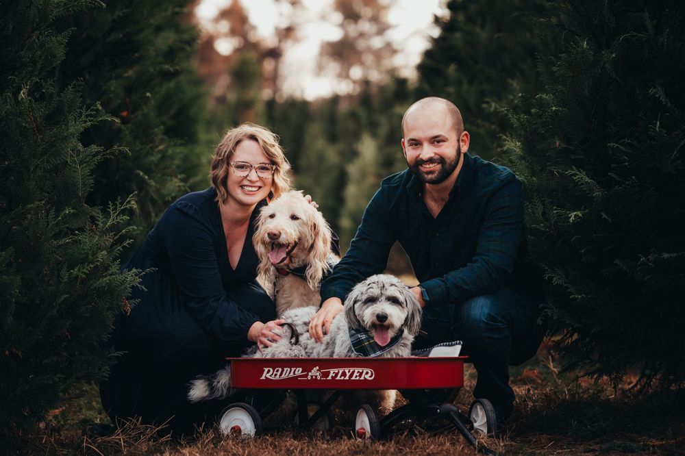 Husband and wife crouch next to red radio flyer wagon where their 2 dogs are sitting in a row of Christmas trees