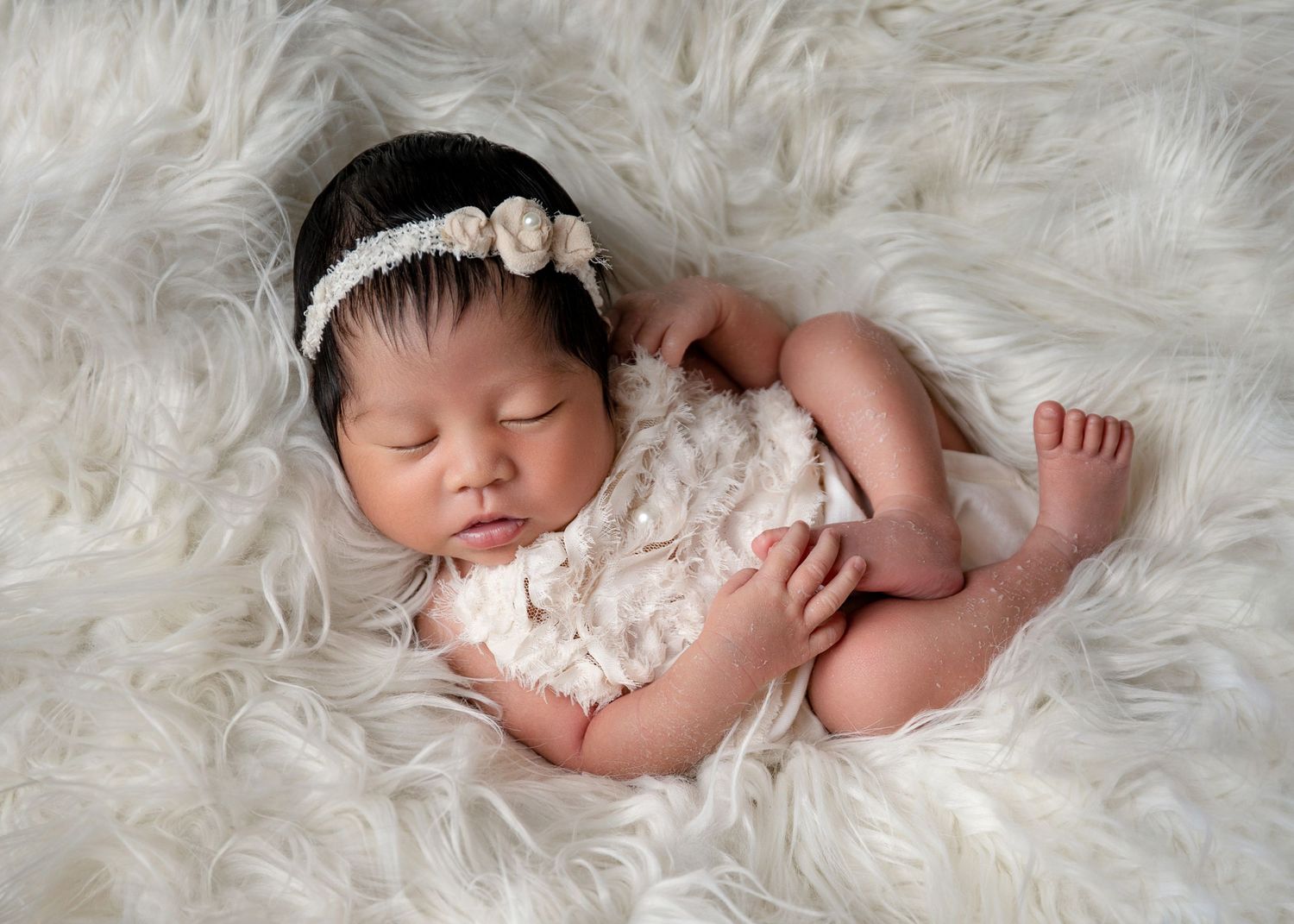 Sleeping newborn wearing lace outfit on soft white fur backdrop.