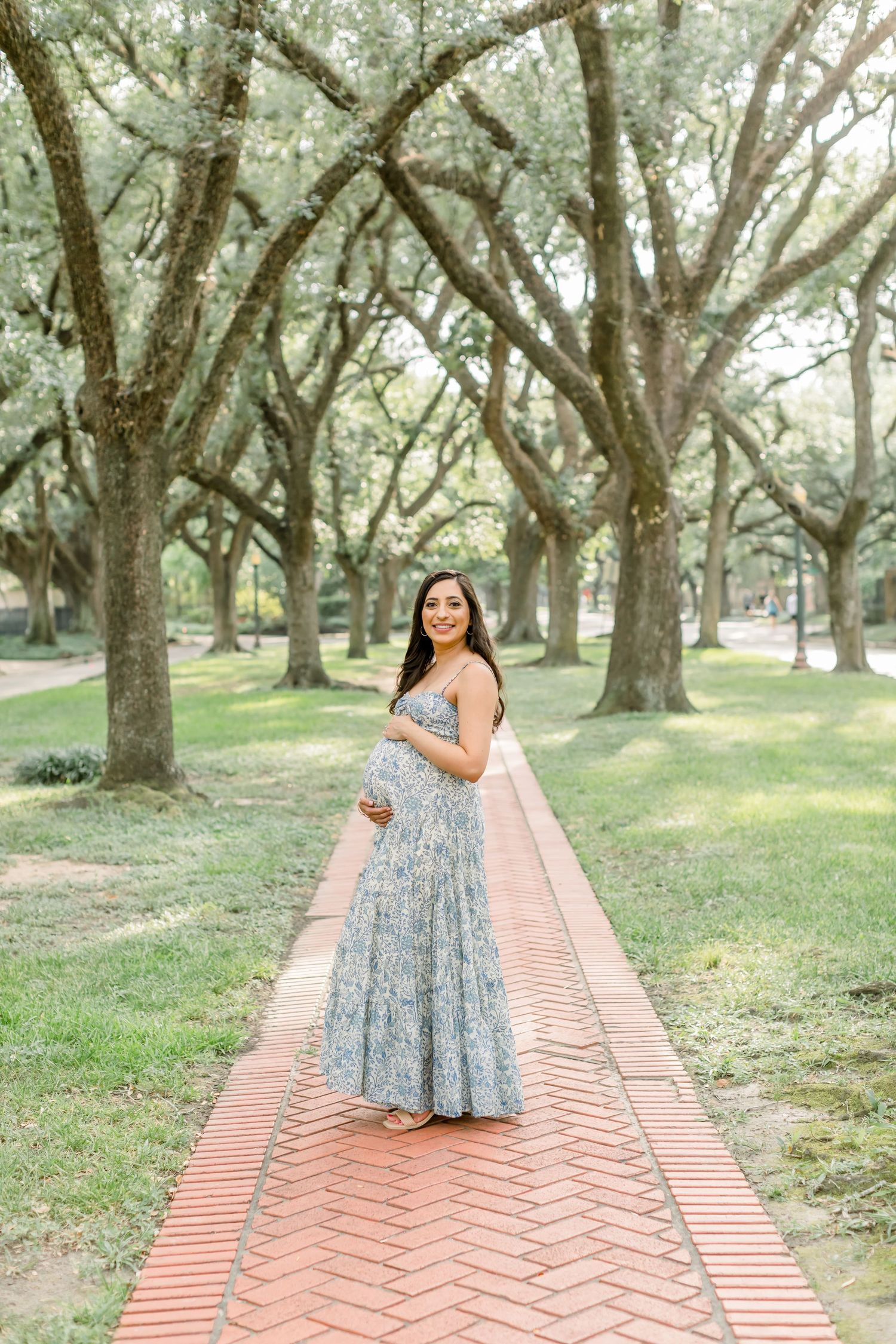 Expecting mother in blue floral maxi dress poses on brick path under oak trees.