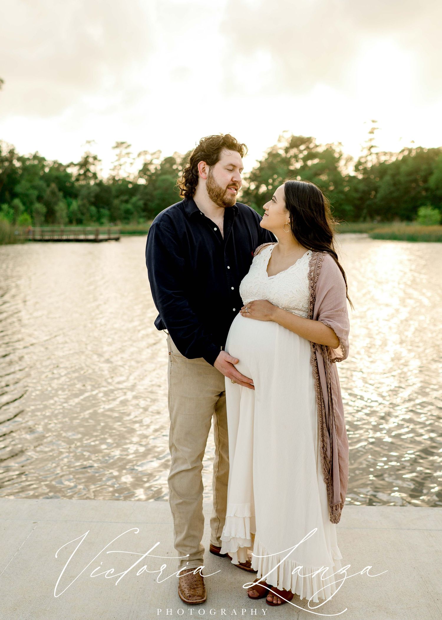 pregnant woman and husban wearing white lace dress posing for a maternity photo session standing behind a lake at sunset