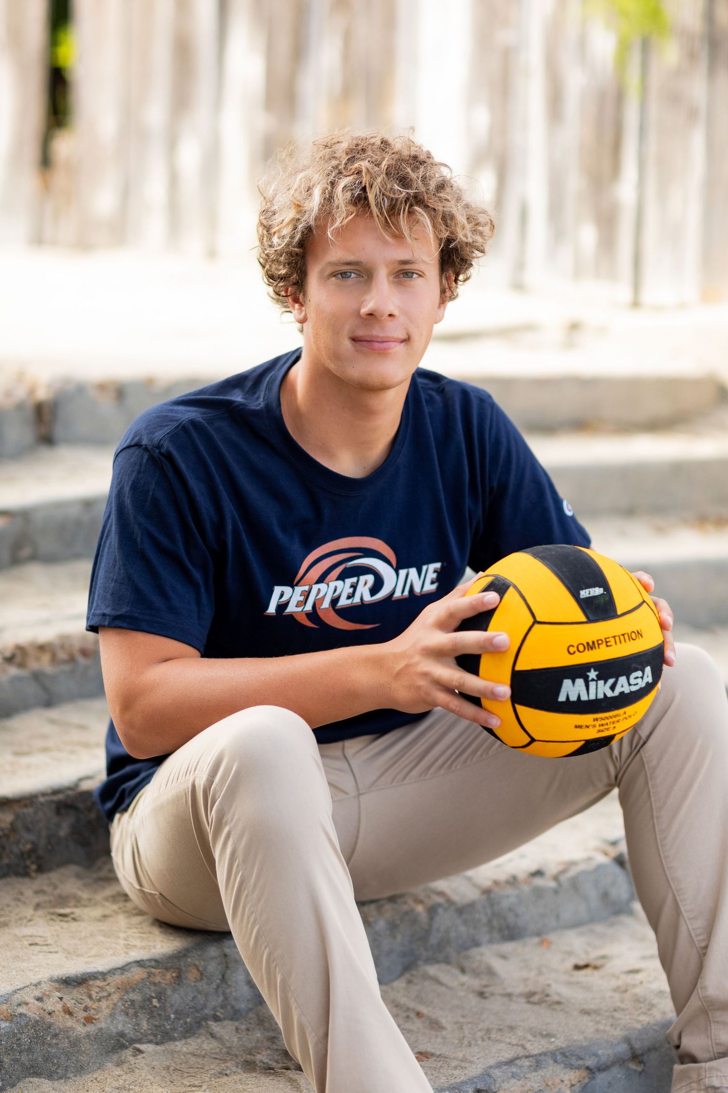 A high school senior athlete in Pepperdine shirt sits casually holding yellow Mikasa water polo ball outdoors.