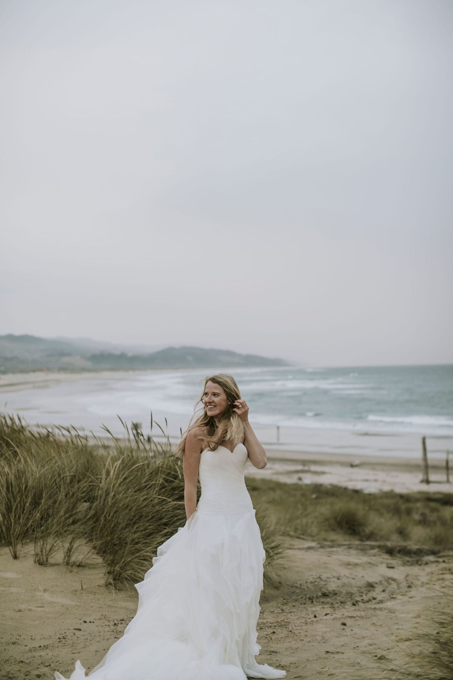Bride posing on the beach in wedding gown with tall grass and ocean behind her