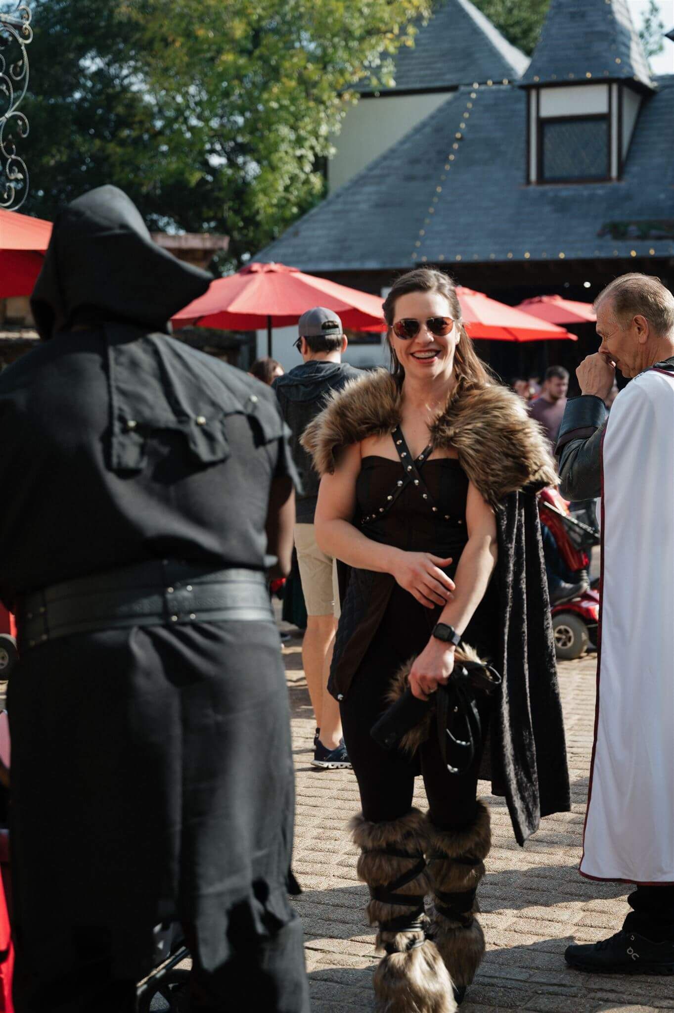 A crowd in medieval costumes mingles at an outdoor renaissance faire with red umbrellas.