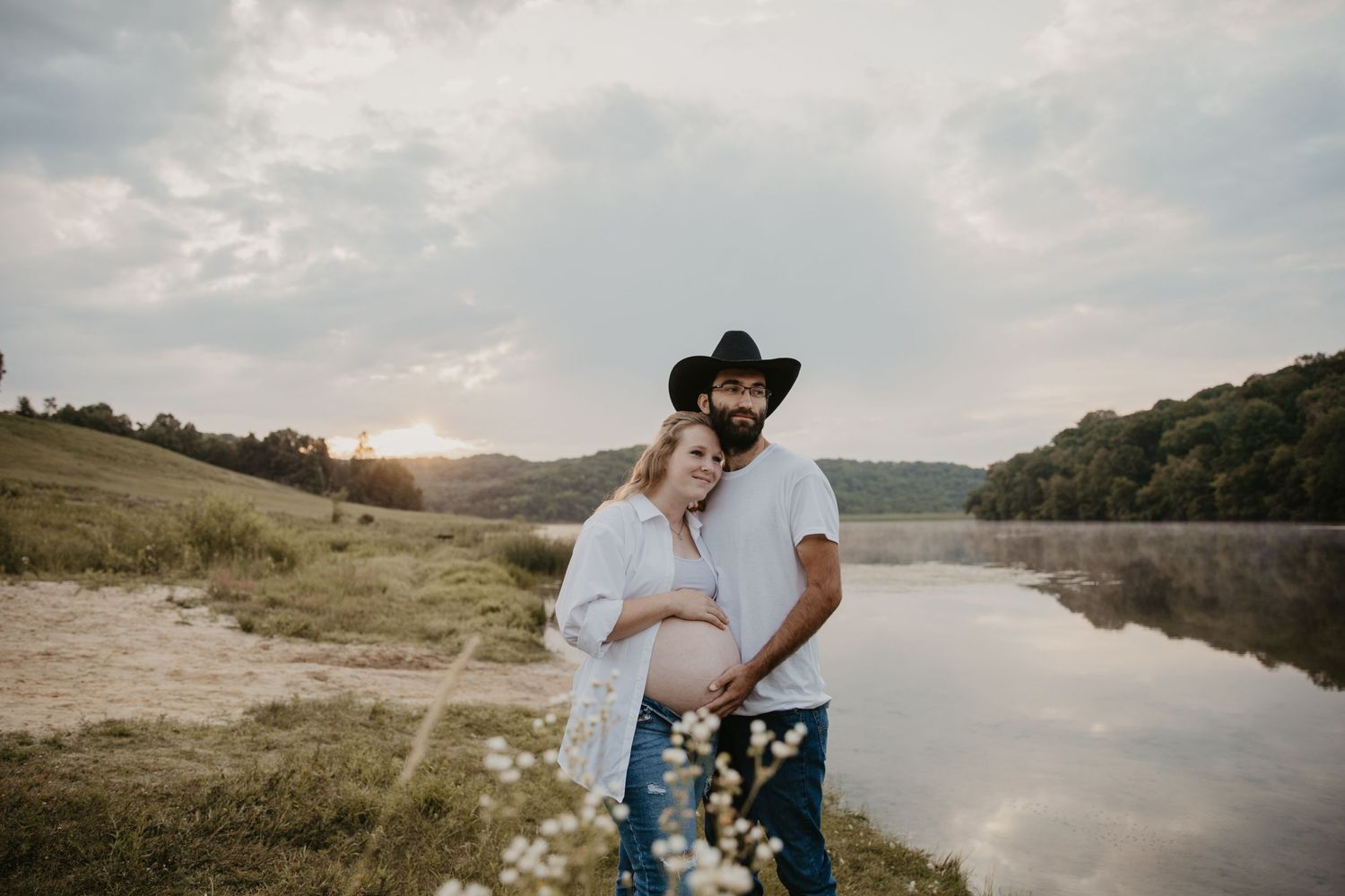couple stands by a lake, embracing each other and facing across the lake, he embraces her big belly with the baby they soon expect