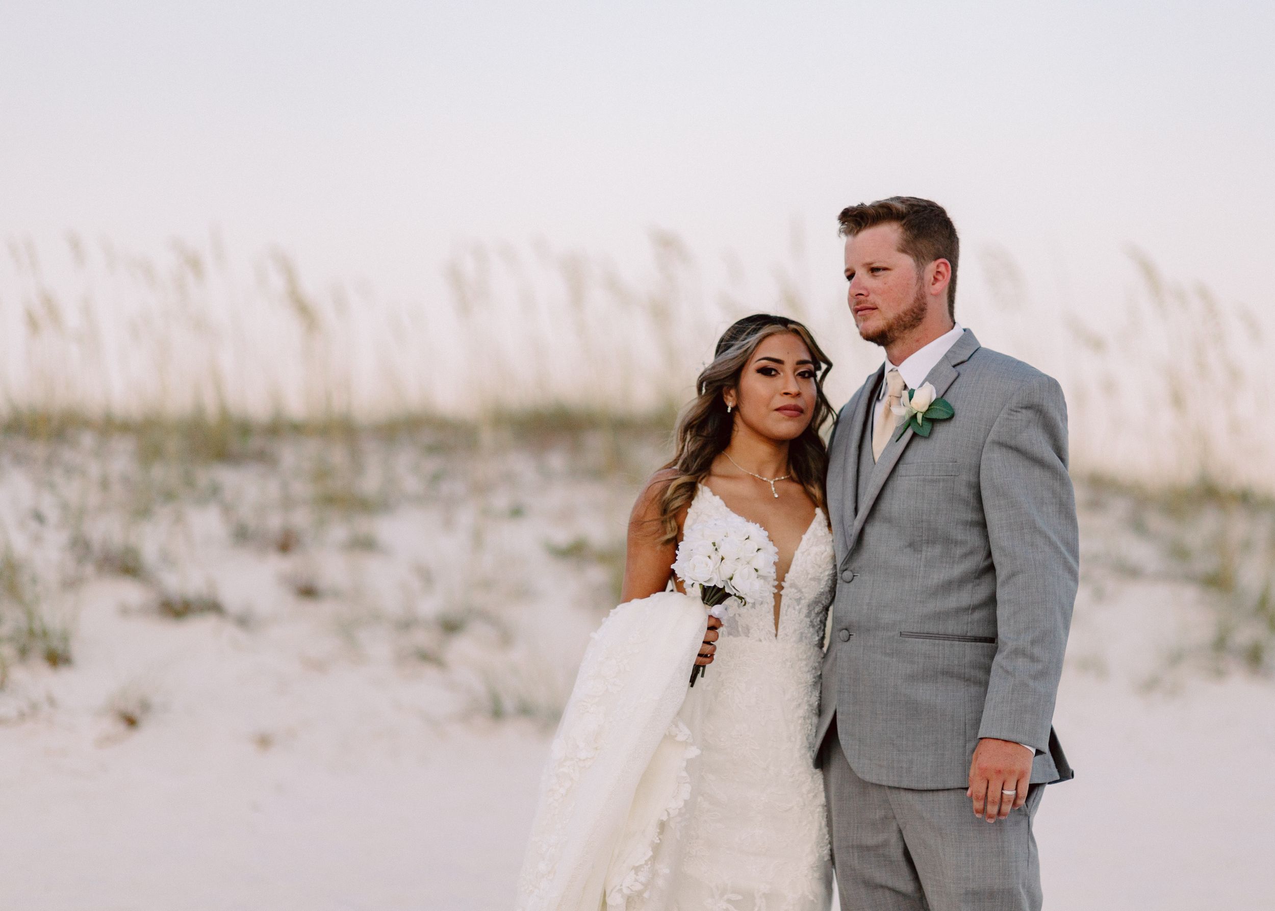 Newlyweds embrace on a white sandy beach during sunset in formal wedding attire.