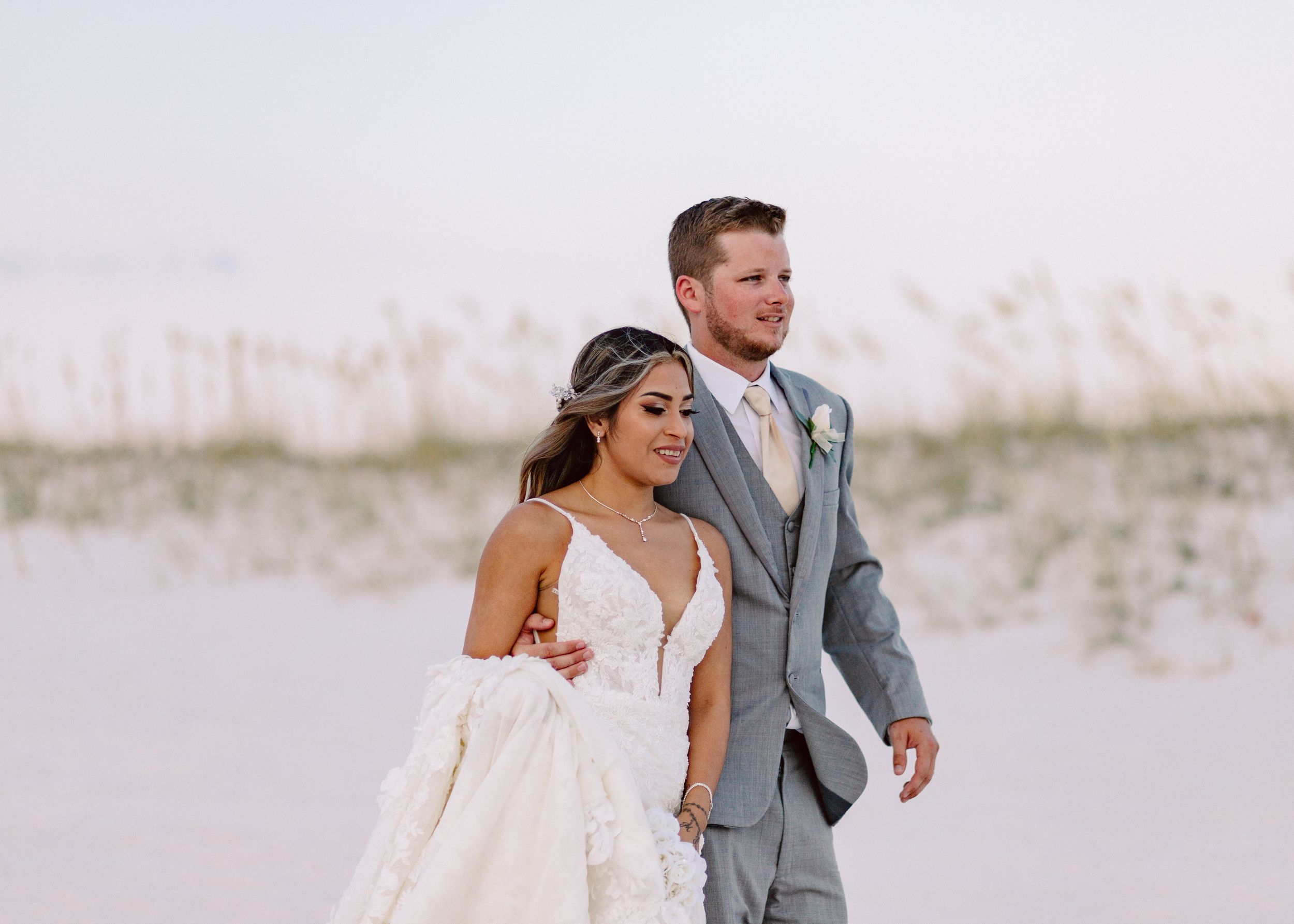 Couple in wedding attire walking along beach dunes during golden hour.