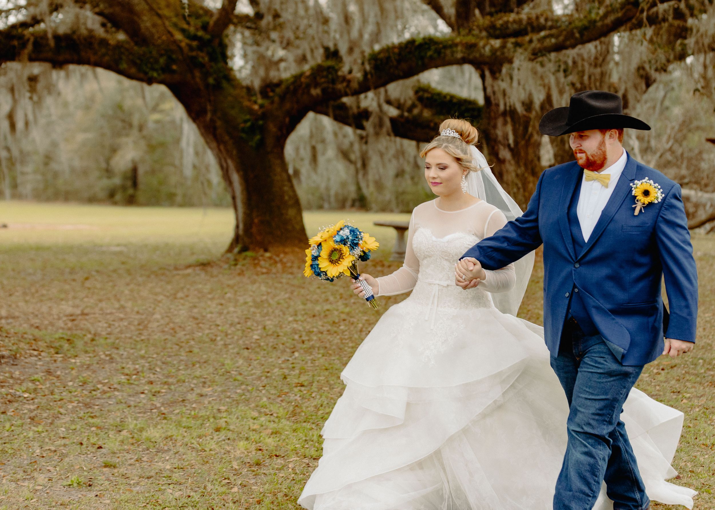 Bride in white gown walks with groom in blue suit under a large oak tree.