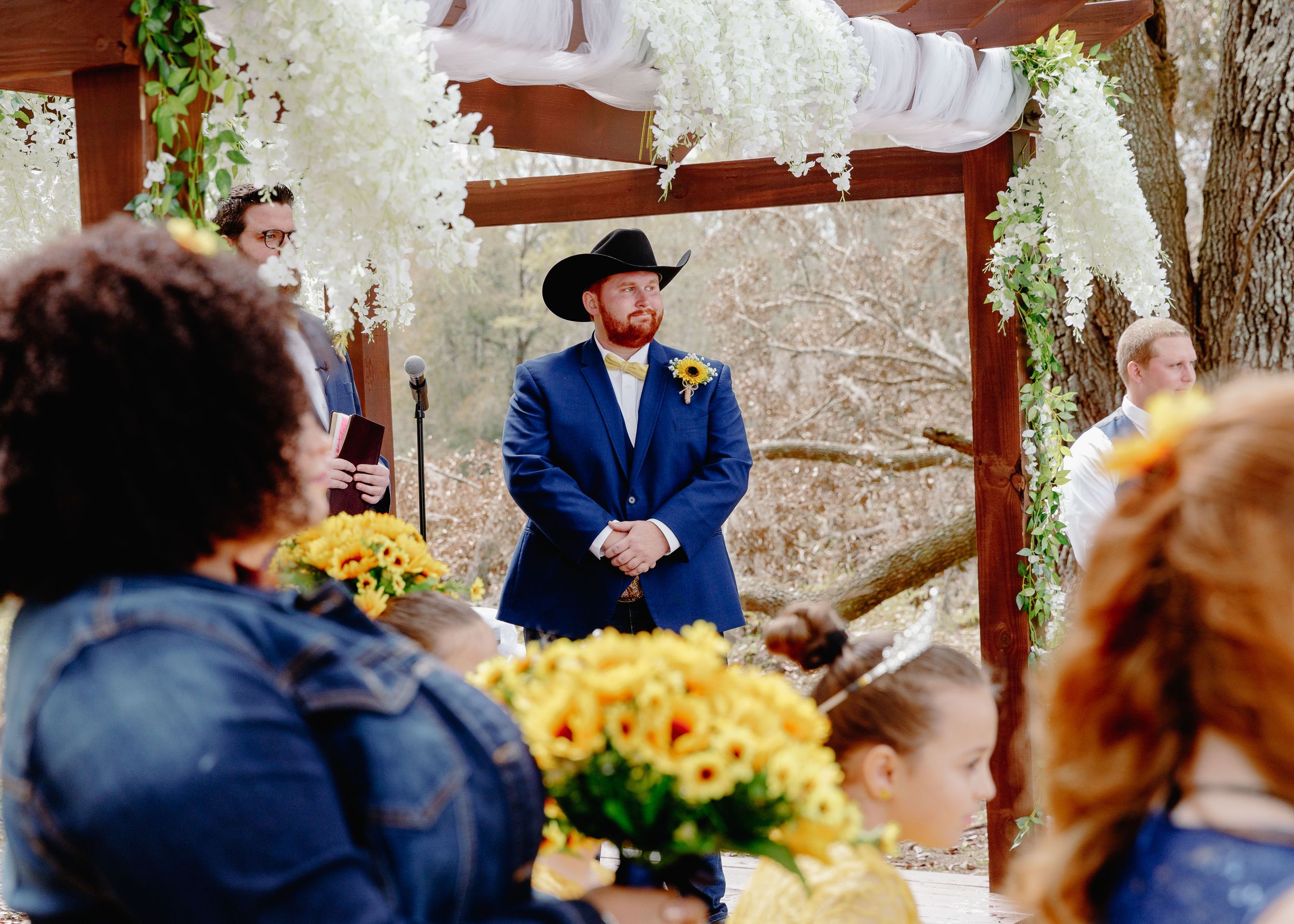 Outdoor wedding ceremony under rustic wooden arbor with yellow floral decorations.