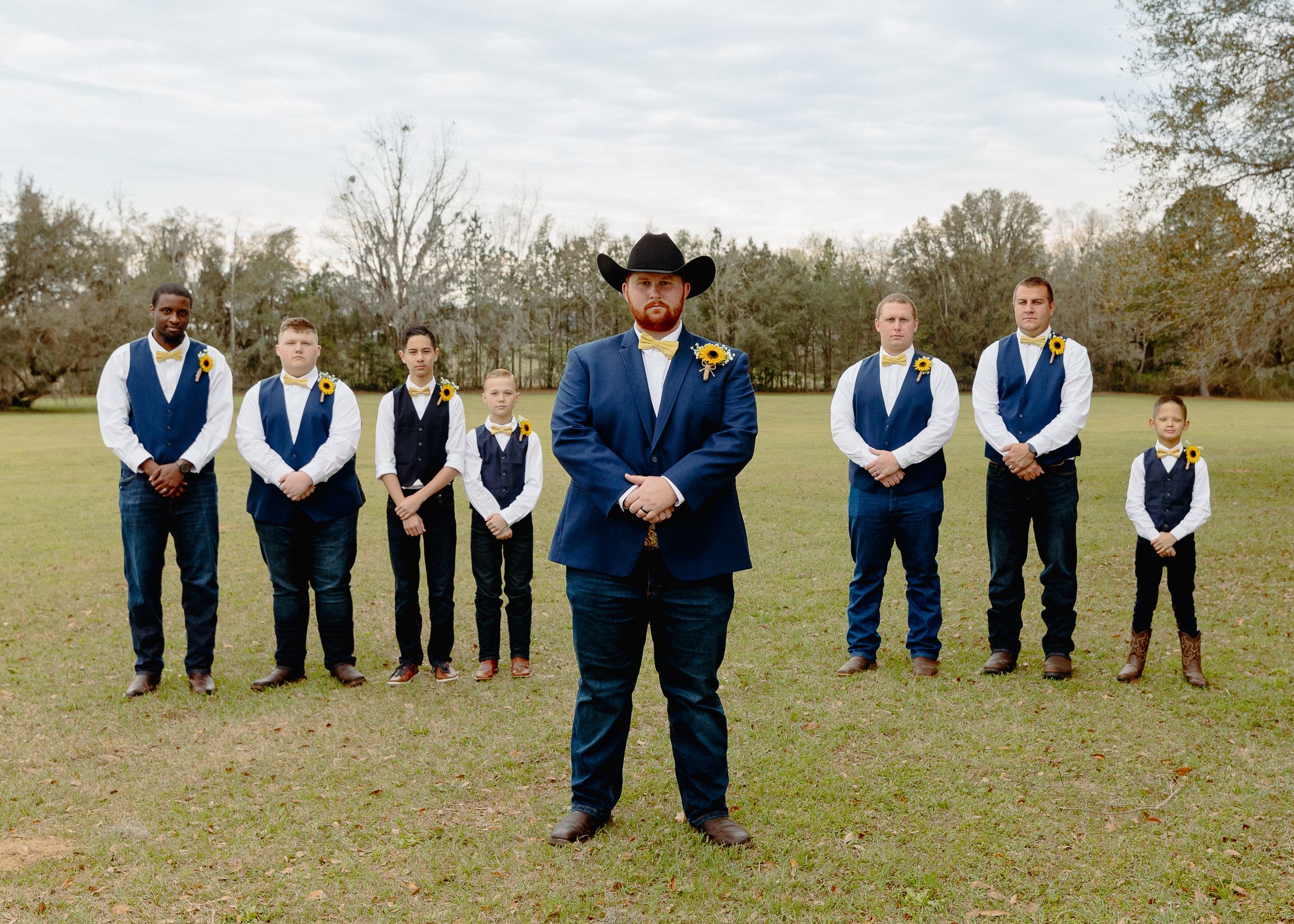 Groomsmen in blue vests stand behind groom in navy suit and black cowboy hat on grassy field.