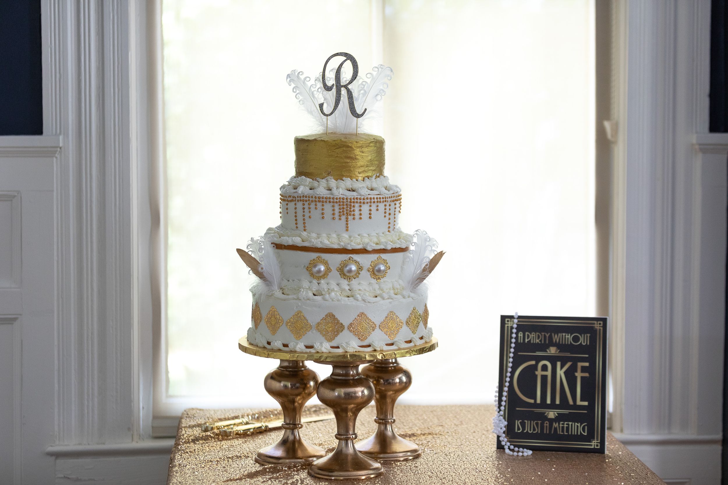 Elegant white and gold wedding cake with monogram topper displayed on ornate gold stand near window.