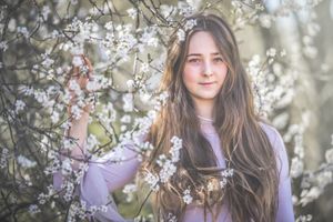 Young woman with long brown hair posing among blooming white cherry blossom tree branches in spring.